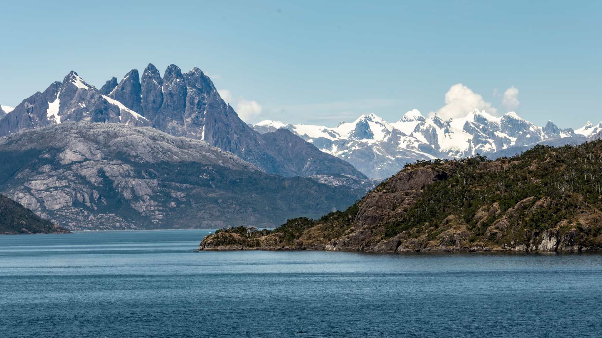 A stunning view of the rugged, mountainous landscape of Chilean Patagonia. The foreground is a wide, dark blue fjord with small ripples on its surface. On the right, a rocky, tree-covered island is visible. In the background, majestic snow-capped mountains and rocky peaks rise against a bright, clear blue sky with a few scattered clouds. The image captures the immense and wild natural beauty of the Patagonian region in South America.