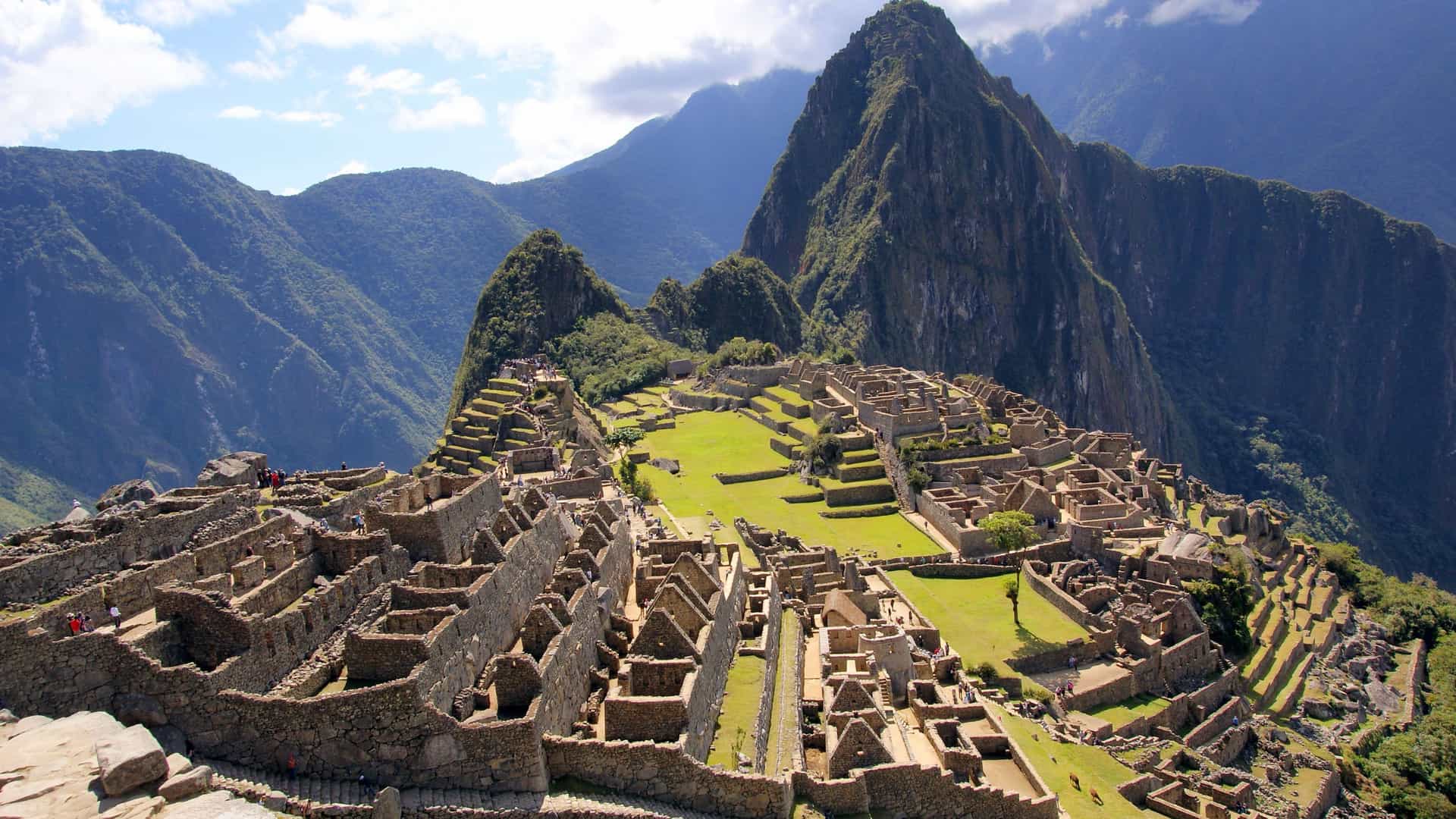 A wide-angle, elevated view of the ancient Inca citadel of Machu Picchu in Peru. The stone ruins of houses, temples, and terraced fields are sprawled across a grassy saddle between two prominent peaks. Behind the main ruins, the iconic triangular peak of Huayna Picchu rises steeply. The entire site is surrounded by the lush, green, and towering Andes Mountains under a partly cloudy, blue sky. The image captures the grandeur and historical significance of this well-preserved South American archaeological site.