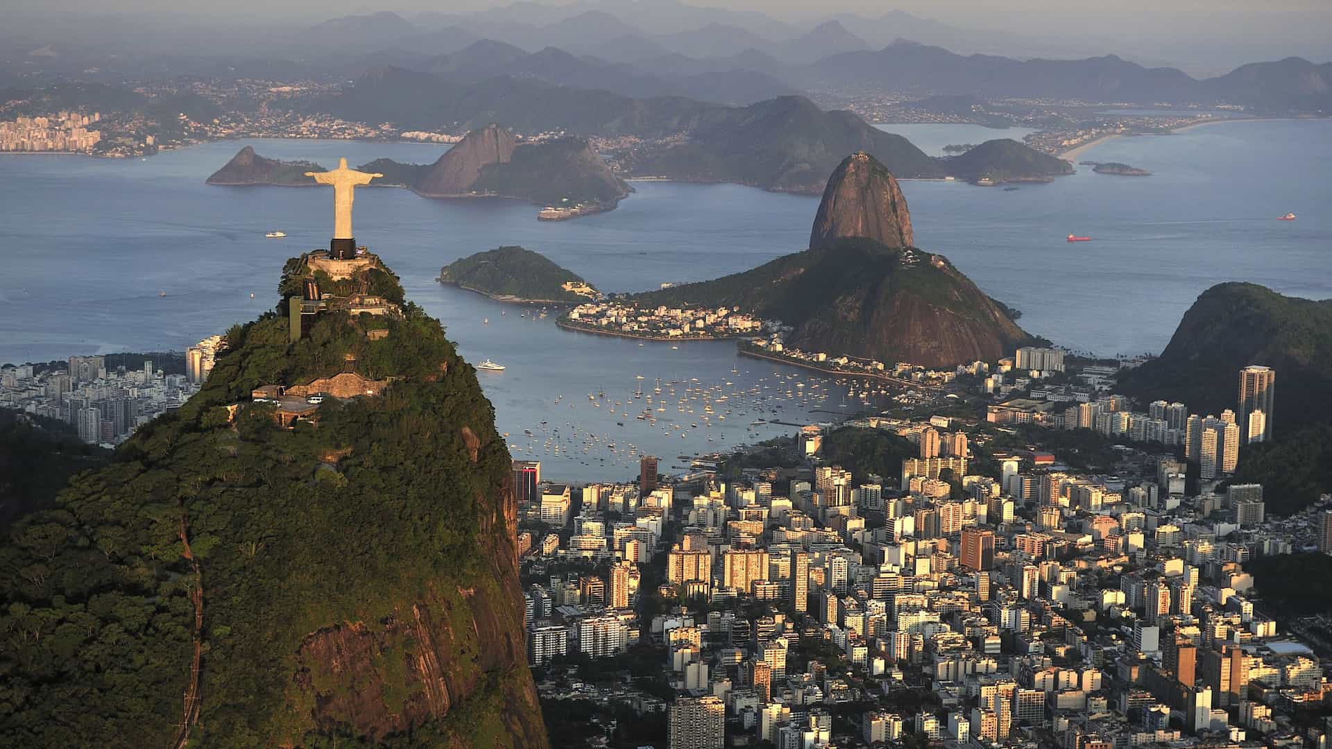 A panoramic aerial view of Rio de Janeiro, Brazil, at sunset. The iconic Christ the Redeemer statue stands with open arms atop a mountain on the left. In the center, Sugarloaf Mountain rises from the water of Guanabara Bay, which is dotted with numerous boats. Below and around the mountains, the city's densely packed buildings are bathed in a warm, golden light. In the distance, more mountains and a hazy coastline stretch to the horizon. The image captures the famous landmarks and stunning coastal geography of this South American city.