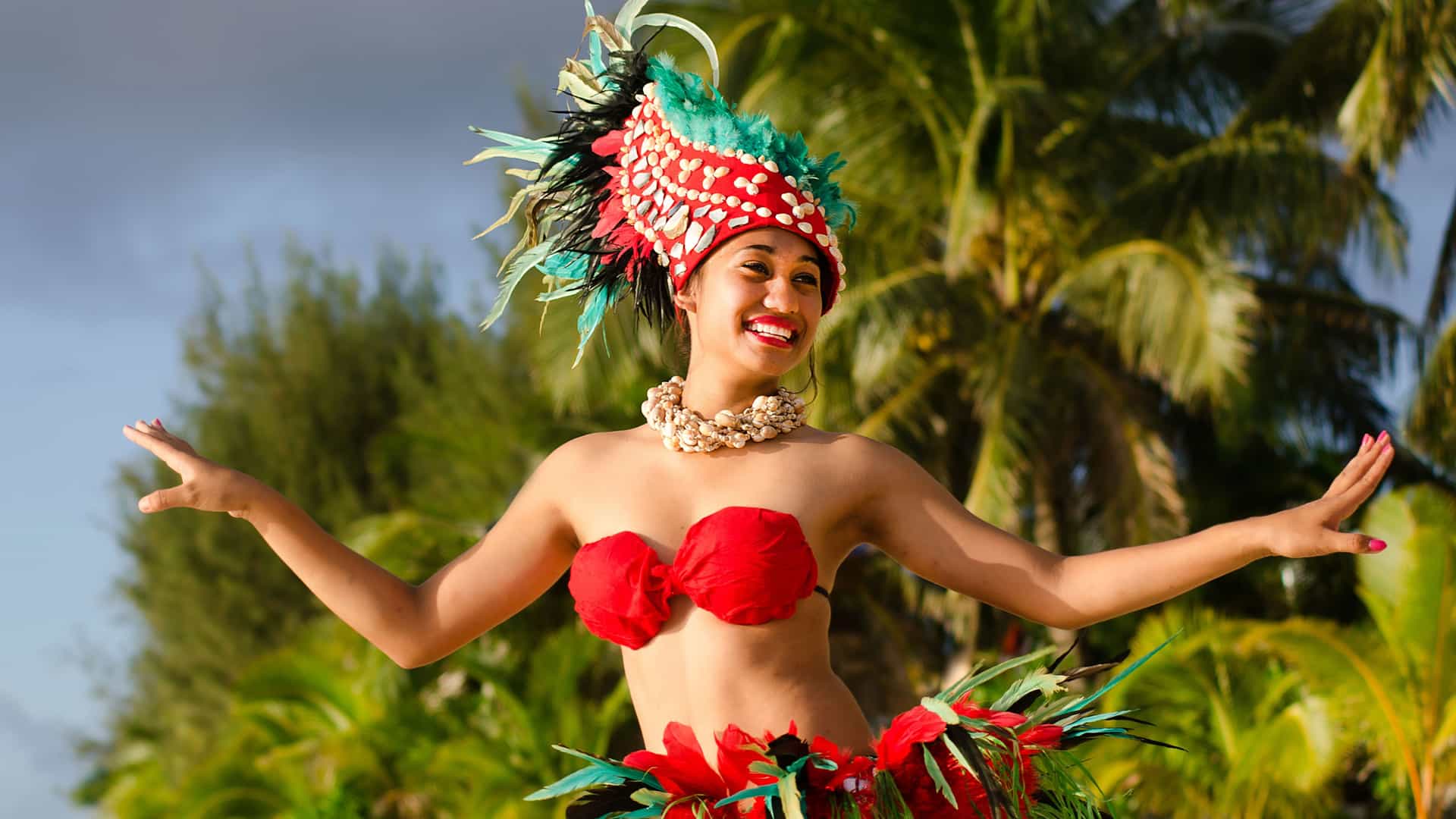 A joyous Polynesian Tahitian dancer performing on a beach in the South Pacific. She wears a vibrant traditional costume, including a red bikini top and a flowing skirt made of red flowers and green leaves. Her elaborate headdress is adorned with red, green, and black feathers, and she wears a necklace of shells. She is smiling broadly with her arms extended, against a backdrop of swaying palm trees and a partly cloudy sky. The image captures the rich cultural traditions and celebratory spirit of the Tahitian region.