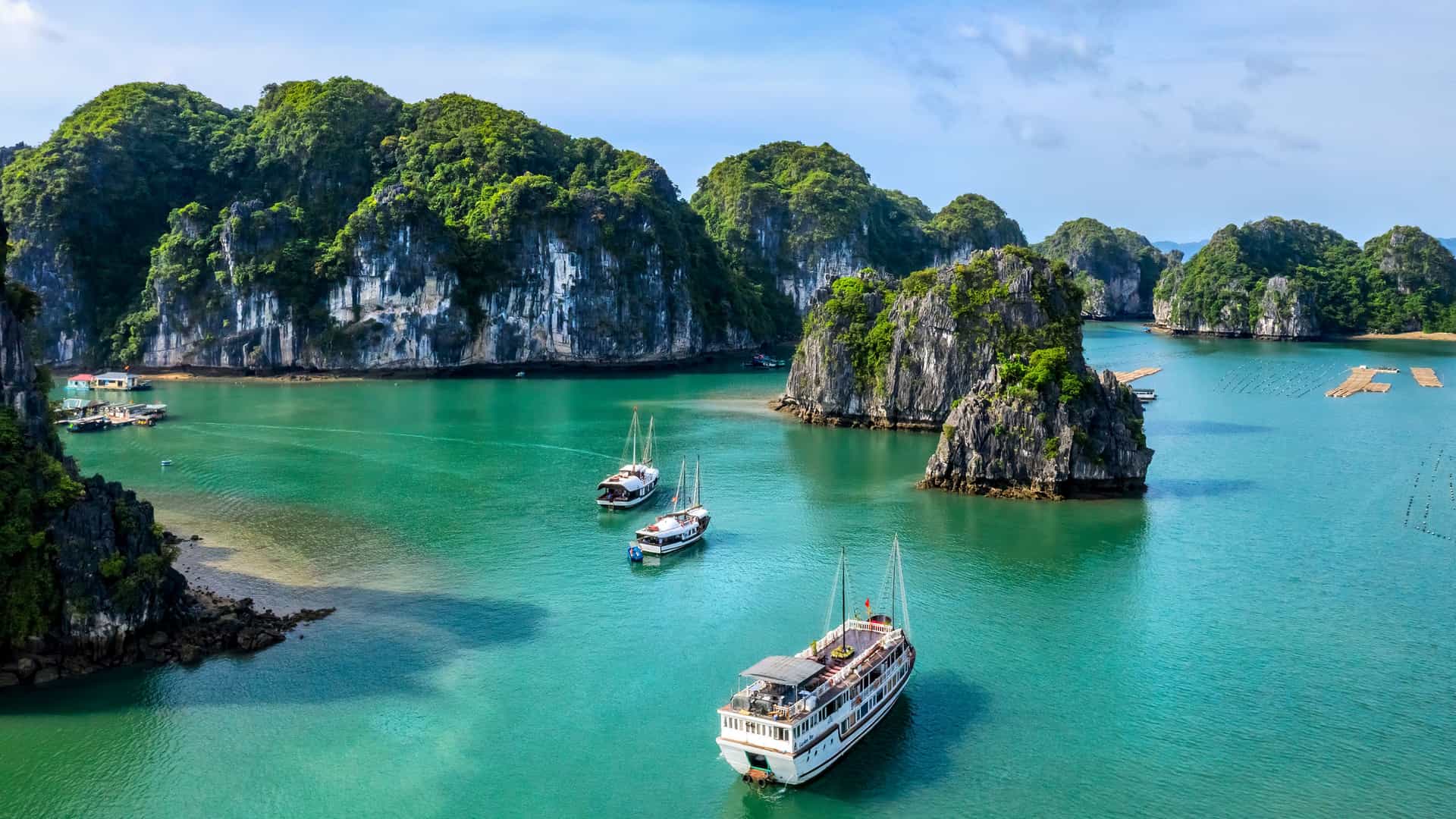 An aerial view of the iconic Ha Long Bay in Vietnam, a UNESCO World Heritage site. The bay is filled with numerous limestone karsts, which are steep, jungle-covered islands of various sizes. The water in the bay is a stunning turquoise-green. Several traditional junk boats and modern cruise ships are visible, either sailing or anchored between the islands. The sky is bright with a few white clouds, and the scene captures the breathtaking natural beauty of this famous Southeast Asian landmark.