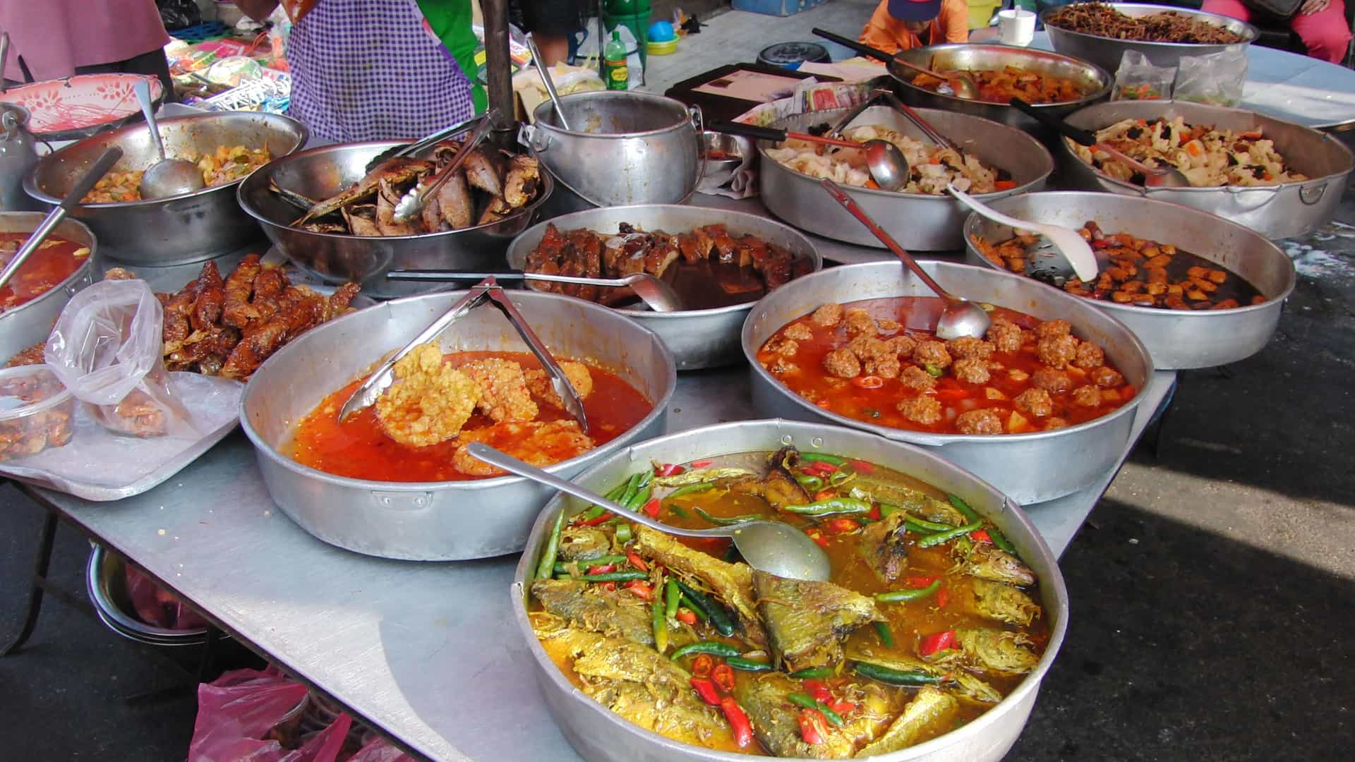 A vibrant display of various traditional Malaysian dishes at a street food market stall in George Town, Penang. The food is presented in large, metal bowls and trays, with different curries, fried fish, meat dishes, and vegetables visible. The food is colorful and appealing, with various sauces and spices. In the background, people are visible browsing the stall. The scene captures the lively and rich street food culture that is a signature part of the Southeast Asian experience in Malaysia.