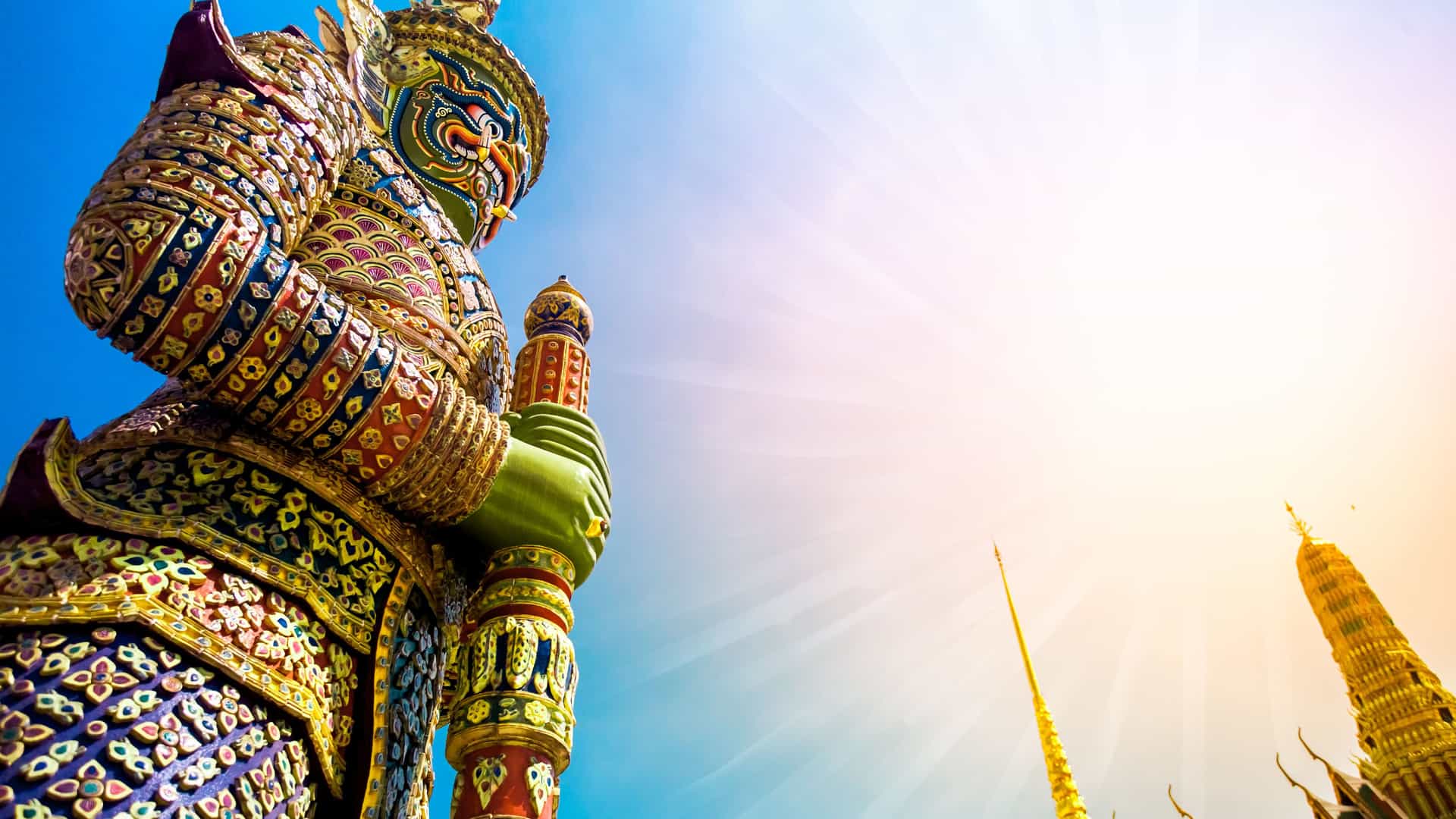 A low-angle, upward shot of a gigantic, colorful statue of a mythical guardian known as a yaksha at Wat Phra Kaew, the Temple of the Emerald Buddha, within the Grand Palace in Bangkok, Thailand. The statue is intricately decorated with mosaic tiles and vibrant colors, and its imposing presence is highlighted against a brilliant blue sky with a bright sunburst. In the background, the golden spires of the temple complex are visible. The image captures the ornate and spiritual artistry found in the historic temples of Southeast Asia.