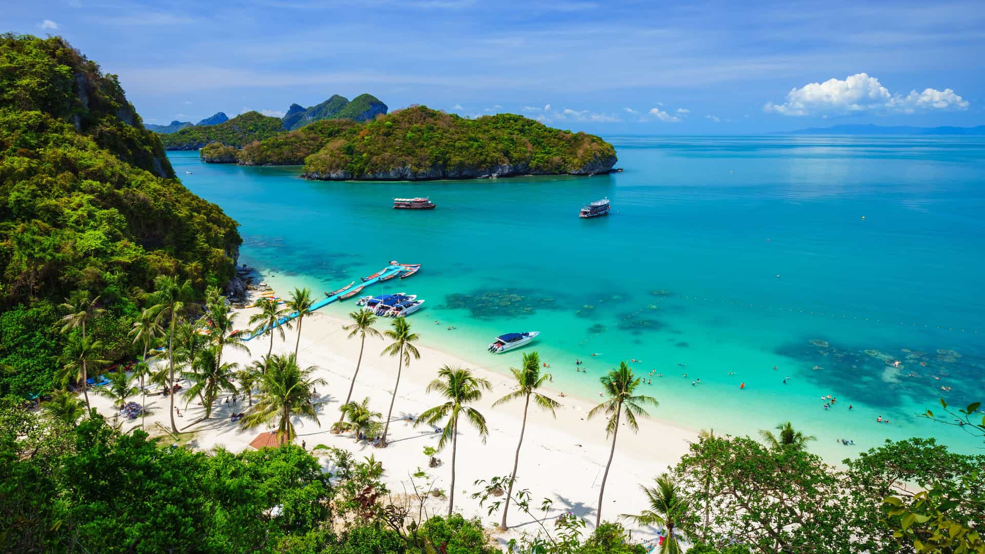 A breathtaking aerial view of a pristine beach on a small island near Koh Samui, Thailand. The white sandy beach is lined with palm trees and extends into a vibrant turquoise bay. Several small boats are moored in the shallow water, and a few people can be seen swimming or relaxing on the beach. In the background, lush, green-covered limestone islands rise from the calm, blue sea under a bright sky with scattered white clouds. The scene captures the idyllic tropical beauty of the Southeast Asian islands of Thailand.