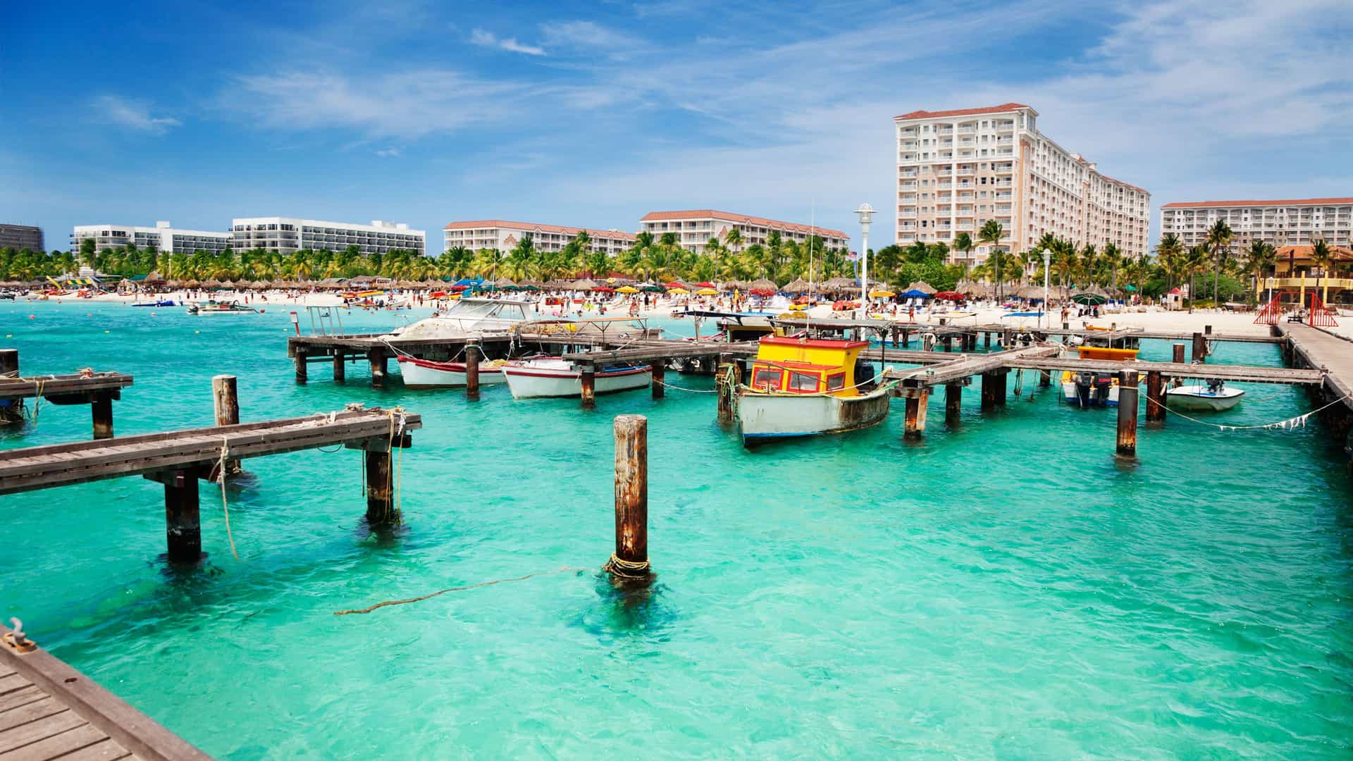 A beautiful view of a resort beach in Aruba, a Southern Caribbean island. The foreground features a series of weathered wooden piers extending into the turquoise waters, with several small boats moored. In the background, a white sandy beach is lined with palm trees and sun loungers. Large, multi-story resort buildings and hotels with balconies and tile roofs stretch along the coastline. The sky is bright blue with scattered white clouds, creating a vibrant and inviting atmosphere that is typical of the Caribbean.