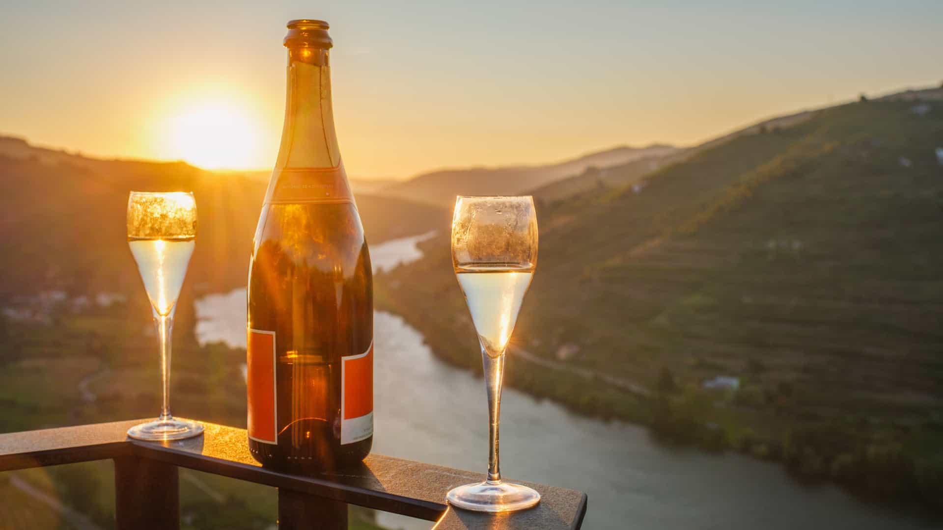 Bottle of sparkling wine with two filled glasses overlooking the Douro River and vineyard hills at sunrise in the Douro Valley, Portugal.