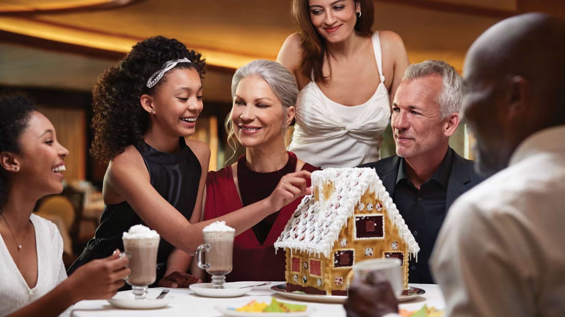 A multigenerational family seated at a formal dining table aboard a cruise ship, surrounding the table as they enjoy decorating a gingerbread houses and drinking hot cocoa.