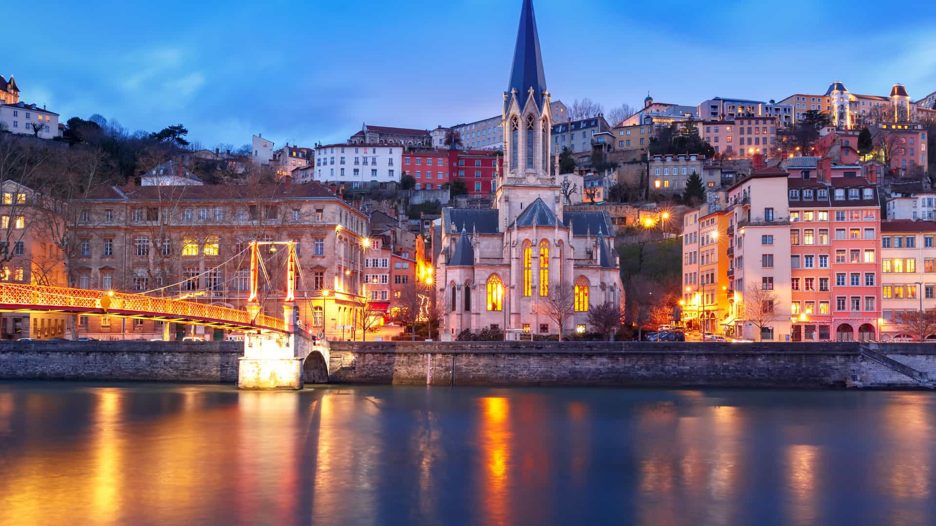 Illuminated church with a tall spire, glowing suspension bridge, and historic architecture along the Saône River in France at dusk on a Tauck river cruise.