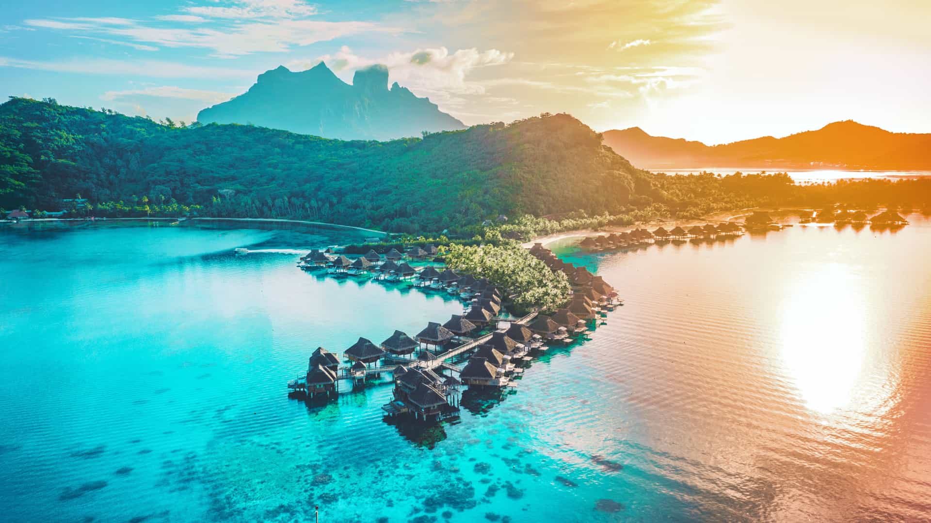 Aerial view of overwater bungalows extending into a turquoise lagoon with lush green mountains on a Tauck South Pacific cruise in Tahiti.