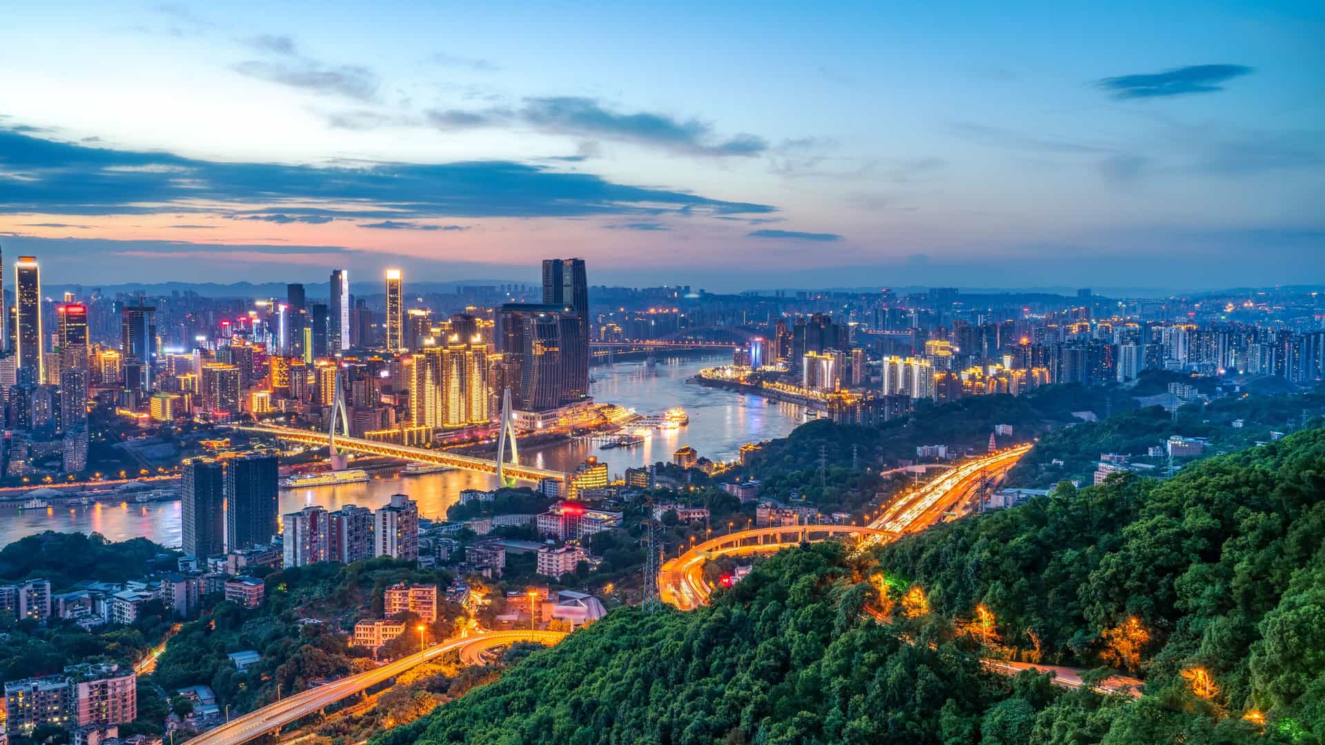 A panoramic dusk view of a modern Chinese city with a river running through it, illuminated skyscrapers, multiple bridges, and winding highways leading in from green hills under a blue‑to‑orange sunset sky.