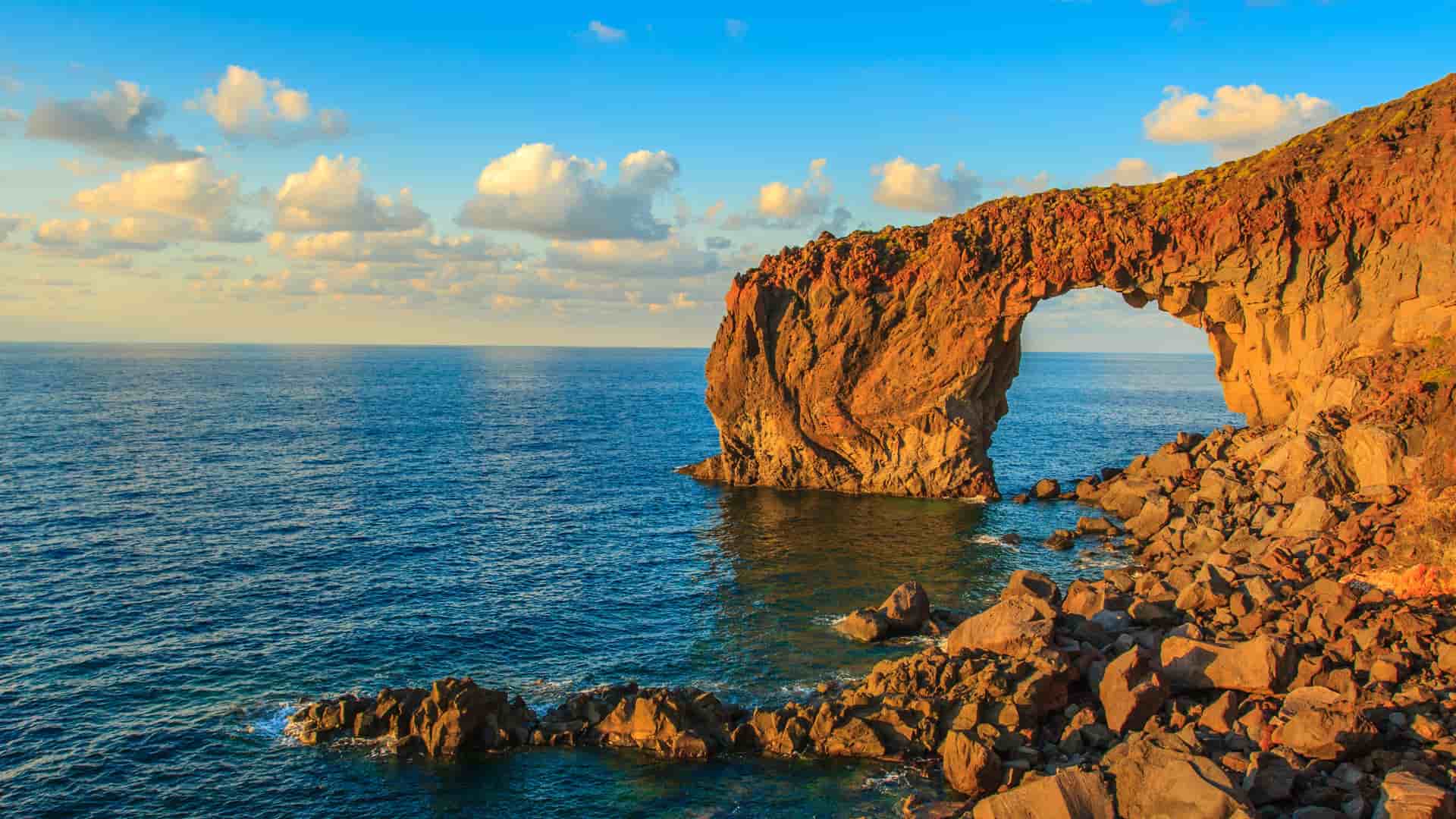 A sunlit coastal rock arch in the Aeolian Islands, Italy, with rugged volcanic cliffs rising above calm blue water under a bright sky, showcasing the dramatic geology of this UNESCO‑listed Mediterranean archipelago.