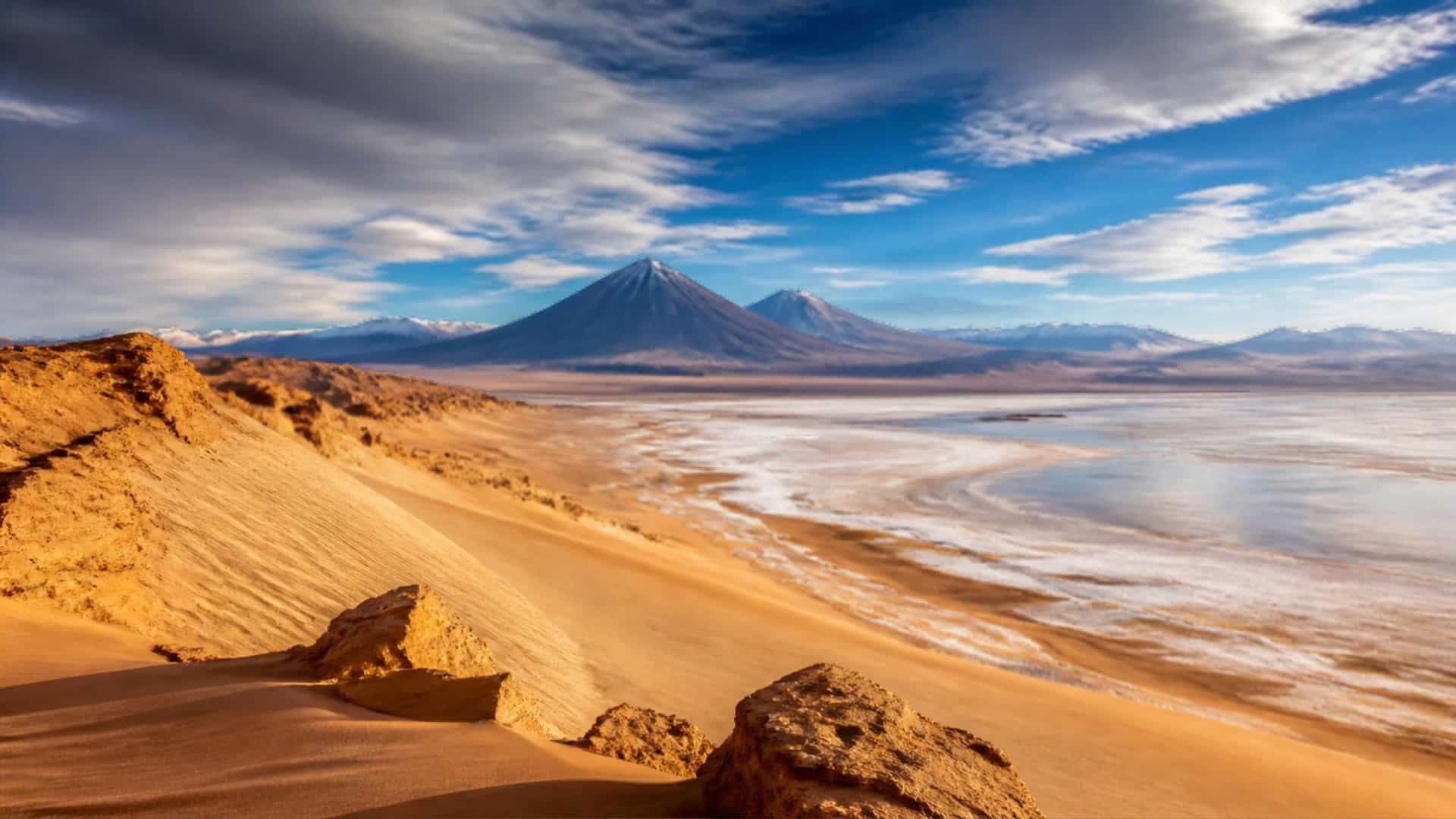 Vast dunes and salt flats of the Atacama Desert in Chile stretch toward distant Andes volcanoes under a dramatic desert sky.