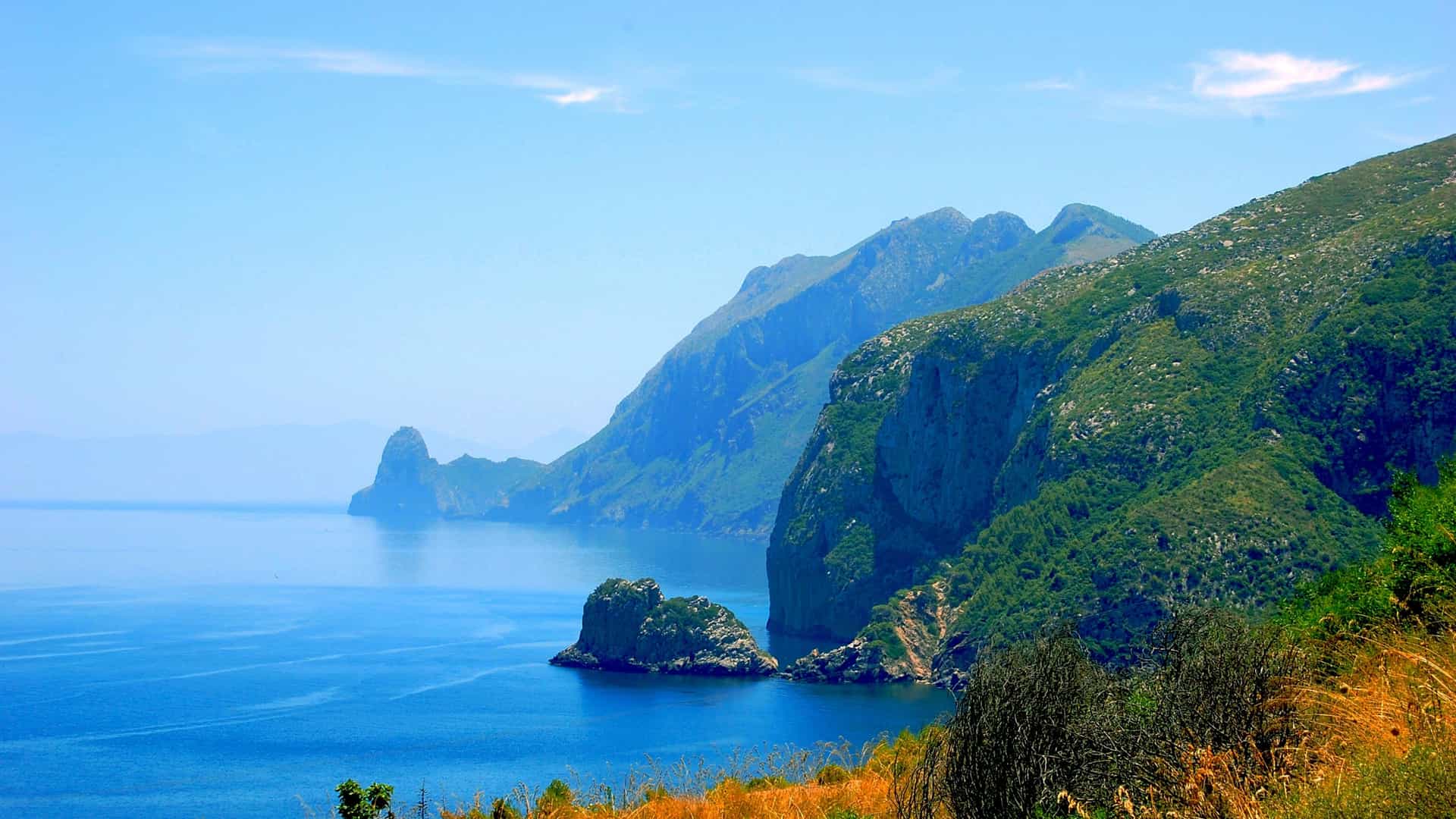 Steep green Bejaia cliffs descending into a deep blue Mediterranean sea, with rocky outcrops and small islands under a clear sky.