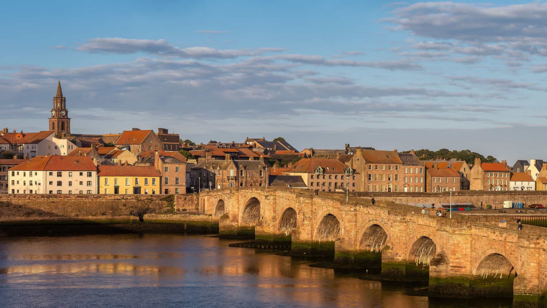 A warm, late‑afternoon view of Berwick‑upon‑Tweed, featuring its historic multi‑arch stone bridge crossing the river, traditional buildings with red‑tiled roofs, and a prominent clock tower glowing in golden light.