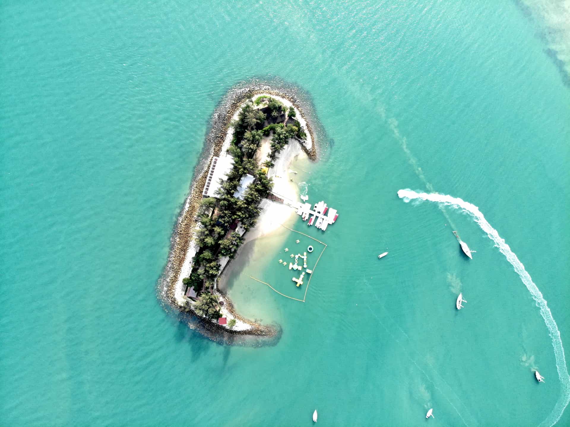 A spectacular high-angle aerial view of a small, privately developed resort islet located off the coast of a tropical cruise destination (frequently cross-referenced with island excursions like those near Bird Island). The teardrop-shaped island features a white sand beach, a dense cluster of green trees, a boat dock, and an inflatable ocean waterpark in its calm lagoon, surrounded by vibrant turquoise water where a speed boat leaves a white wake.