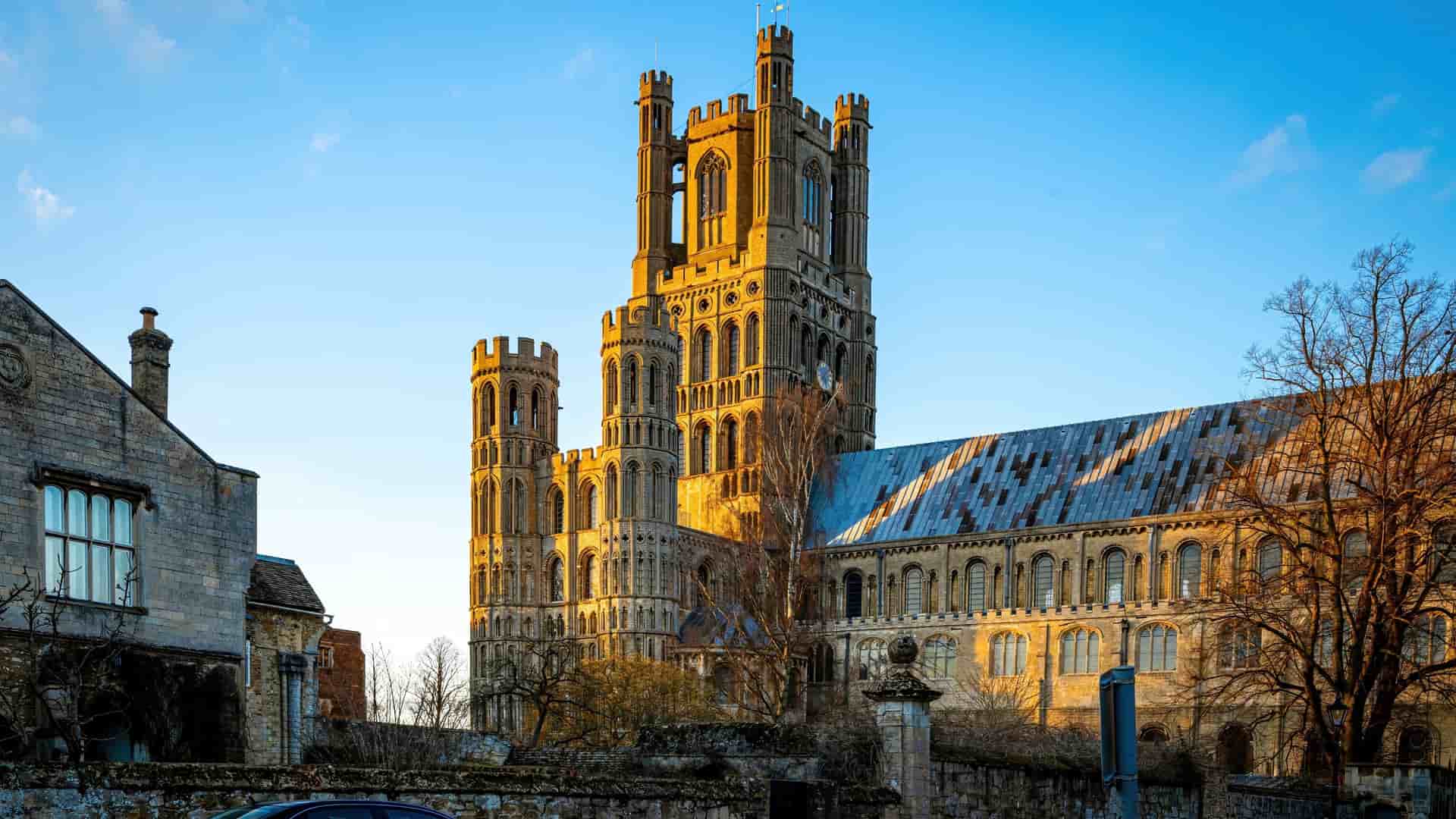 A stunning side-angle view of the historic Ely Cathedral near Cambridge, England—a magnificent landmark and popular regional shore excursion destination. The image captures the cathedral's grand Romanesque and Gothic stone architecture, highlighted by its towering West Tower glowing in the warm light of a late afternoon sun against a clear blue sky, with older brick buildings and trees framing the foreground.