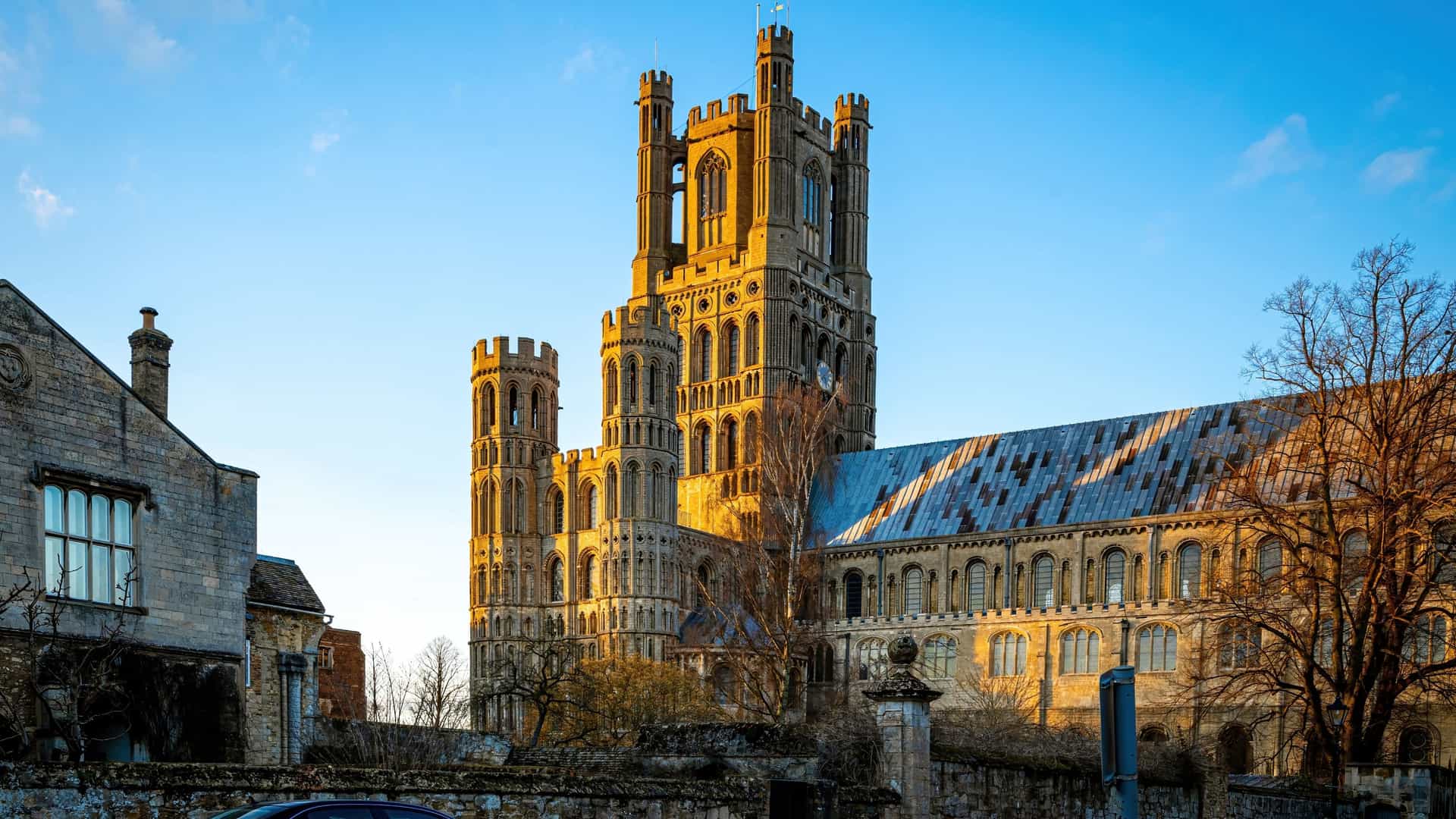 A stunning side-angle view of the historic Ely Cathedral near Cambridge, England—a magnificent landmark and popular regional shore excursion destination. The image captures the cathedral's grand Romanesque and Gothic stone architecture, highlighted by its towering West Tower glowing in the warm light of a late afternoon sun against a clear blue sky, with older brick buildings and trees framing the foreground.
