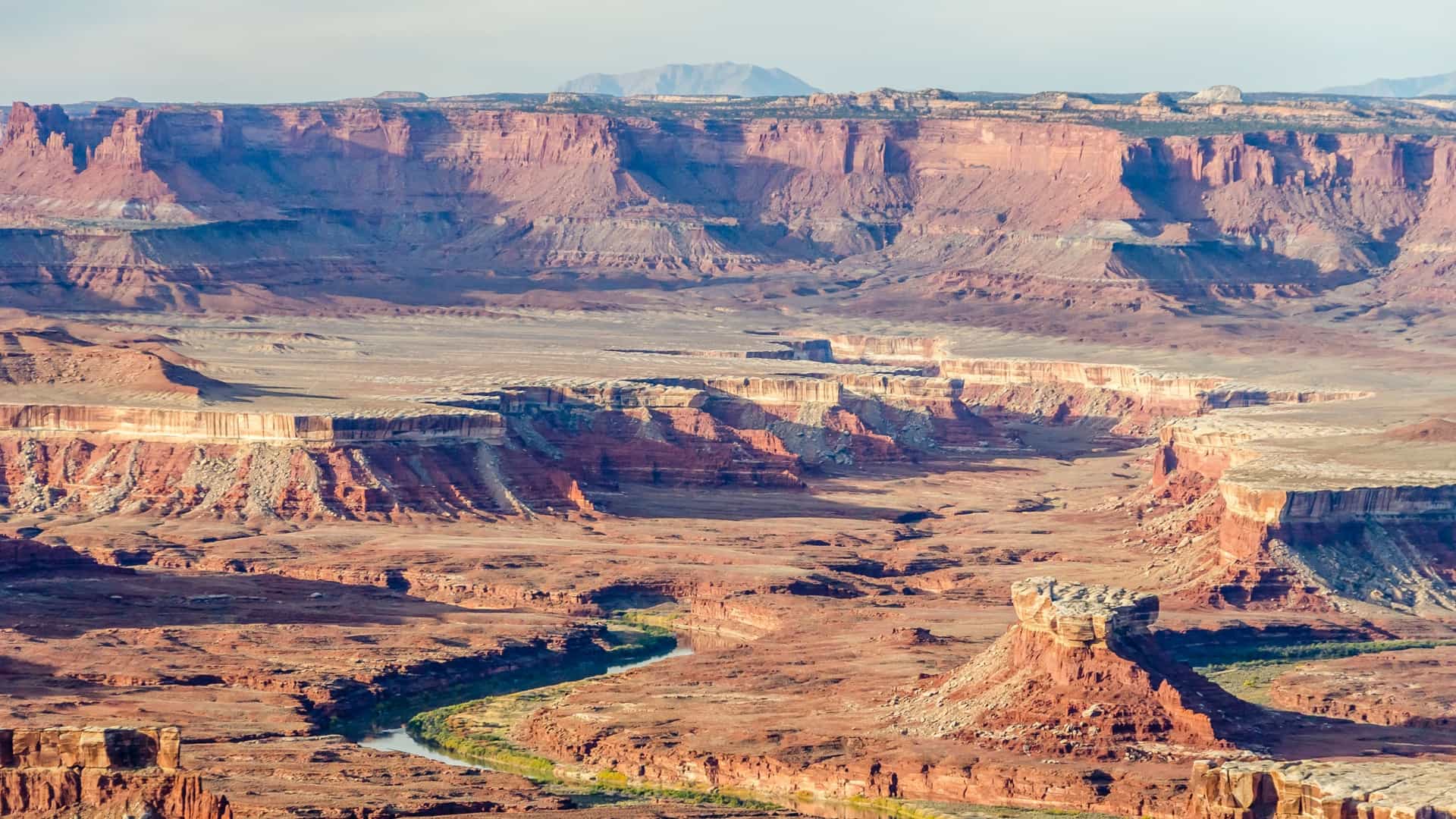 A breathtaking high-angle view overlooking the vast, multi-layered red rock canyons of Canyonlands National Park in Utah, USA—a premier overland shore excursion destination. The image captures the deep, sweeping expanse of the Green River cutting a winding path through the rugged desert floor, flanked by massive flat-topped mesas, sheer cliffs, and unique rock towers under a soft, hazy sky.