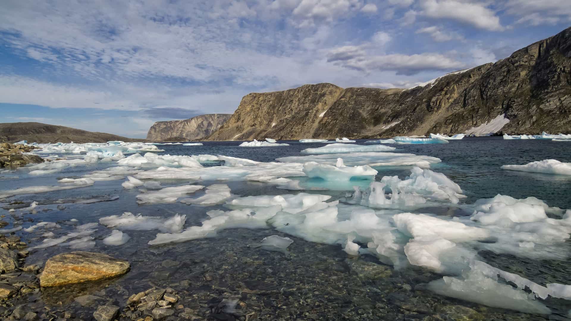 A rocky Arctic shoreline at Cape Mercy with floating ice chunks and small icebergs drifting in cold blue water, backed by steep snow‑patched cliffs under a partly cloudy sky.