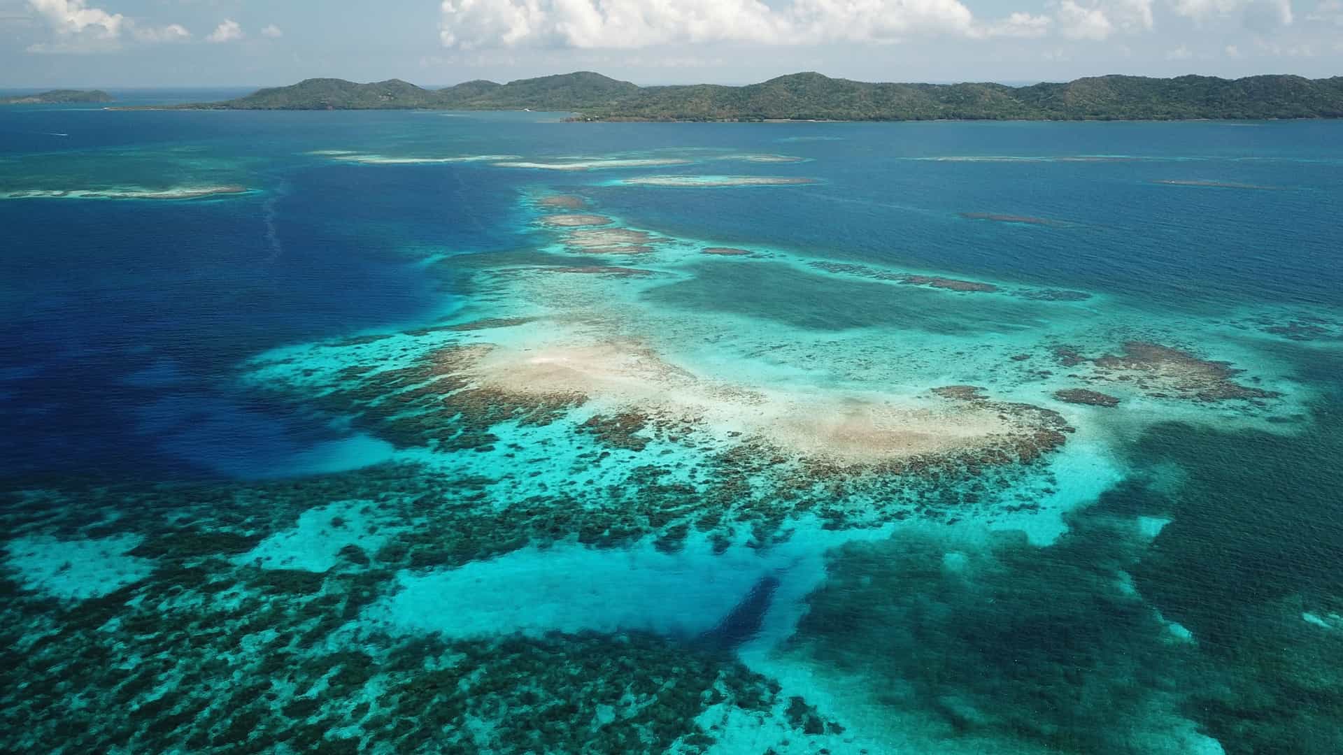 An aerial view of Cayos Cochinos showing a bright turquoise coral reef surrounded by deep blue water, with green hilly islands in the background under a partly cloudy tropical sky.