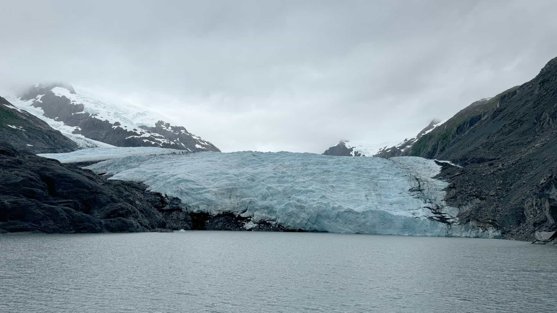 A striking landscape view of a massive, textured tidewater glacier terminating into a calm gray bay in Alaska, USA—a premier destination for glacier viewing boat tours and wilderness shore excursions (frequently accessed via the Kenai Fjords region near the Chiswell Islands). The image captures the dense blue ice of the glacial face nestled tightly between steep, rugged dark rock mountainsides under an overcast sky.
