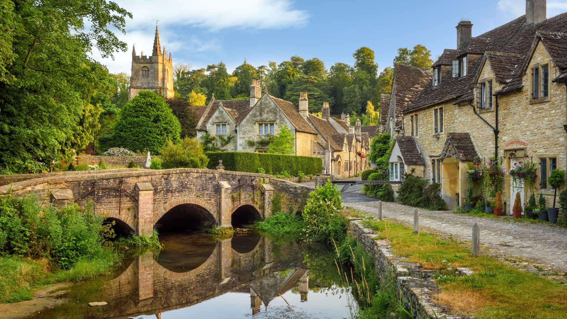 A postcard-perfect, eye-level view of the historic village of Castle Combe in the Cotswolds, England—a quintessential British countryside shore excursion destination. The image features a classic three-arched stone bridge spanning a calm, reflective river, leading the eye past honey-colored stone cottages with slate roofs and climbing ivy, with a medieval church tower rising above the lush green trees in the background under a soft blue sky.