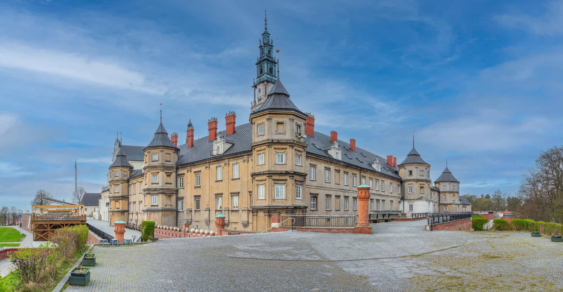 A grand, side-angle view of the historic Jasna Góra Monastery in Częstochowa, Poland—a world-famous shrine and premier cultural shore excursion destination. The image highlights the monastery's sprawling baroque stone complex, featuring defensive brick bastions, tiered corner towers with conical roofs, and a tall, decorative green-capped spire rising into a cloudy blue sky.