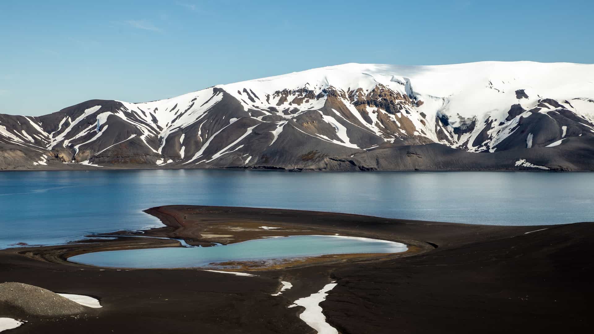 A volcanic crater lake on Deception Island surrounded by black ash slopes, turquoise pools, and snow‑capped mountains under a bright Antarctic sky.