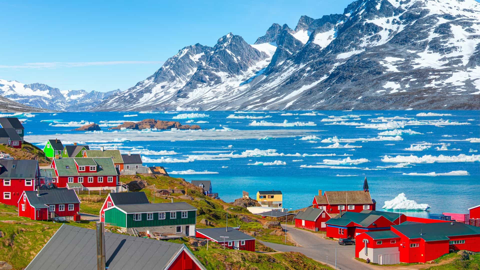 A vibrant, high-angle view of the colorful coastal village of Tasiilaq on the east coast of Greenland—a spectacular Arctic cruise destination. The image showcases a collection of bright red, green, and blue Nordic houses terraced along a grassy green hillside, overlooking a brilliant blue bay filled with drifting ice floes and icebergs, backed by massive, snow-capped jagged mountain peaks under a clear blue sky.