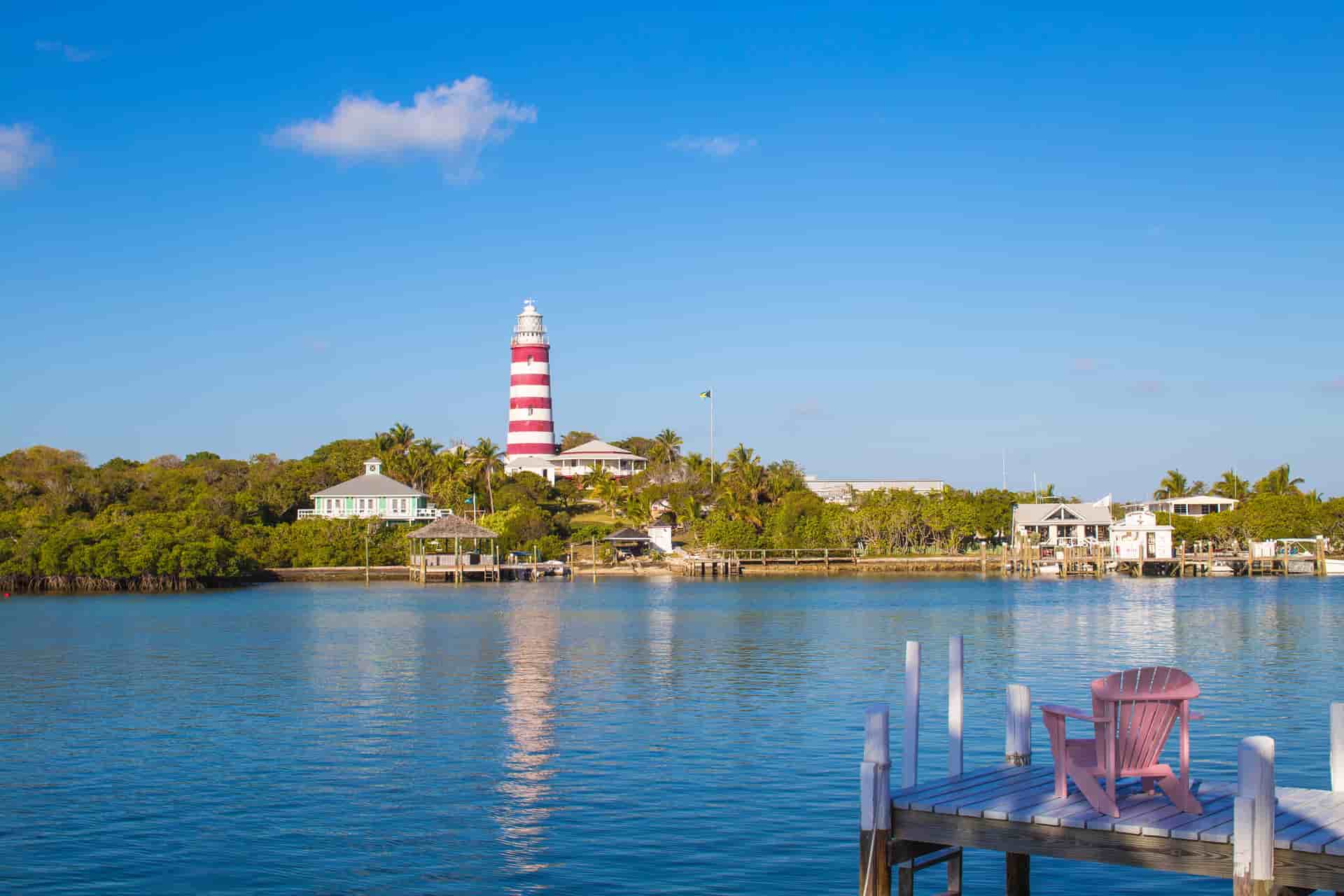 The image highlights the iconic red-and-white candy-striped Elbow Reef Lighthouse rising above lush green trees and coastal homes, with a pink wooden Adirondack chair sitting on a white boat dock in the immediate right foreground under a clear blue sky.