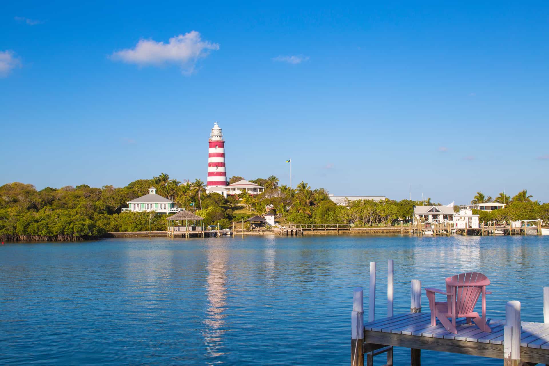 The image highlights the iconic red-and-white candy-striped Elbow Reef Lighthouse rising above lush green trees and coastal homes, with a pink wooden Adirondack chair sitting on a white boat dock in the immediate right foreground under a clear blue sky.