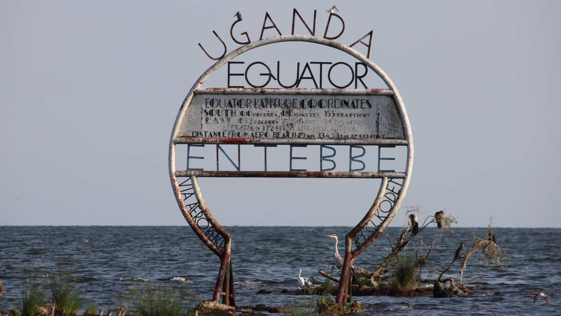 The Entebbe Equator marker standing in Lake Victoria, with rusted metal rings, perched birds, and calm water surrounding the landmark.