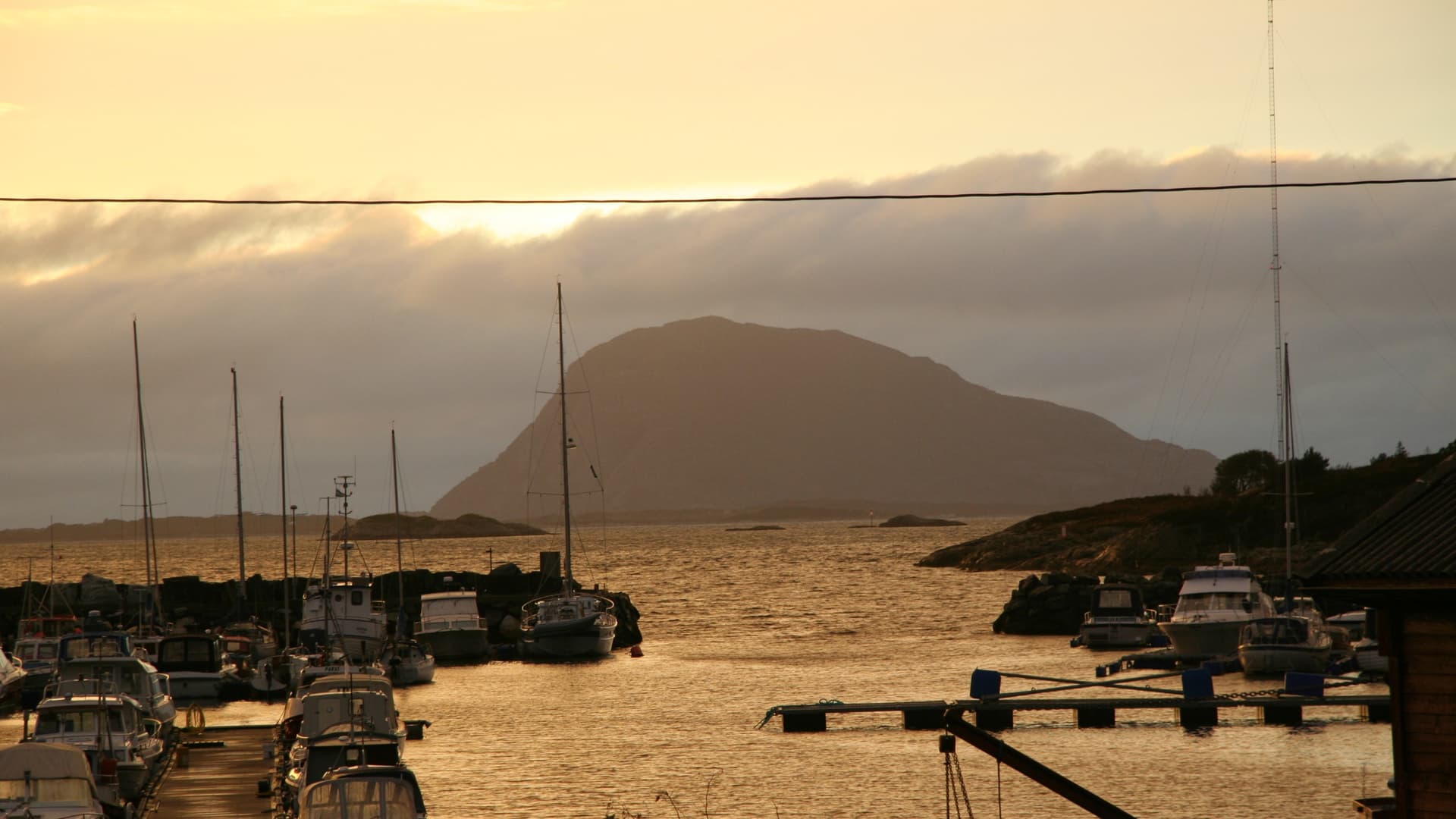 Golden sunset over Florø's harbor in Norway, with fishing boats and sailboats moored in the calm water and forested coastal hills rising behind.