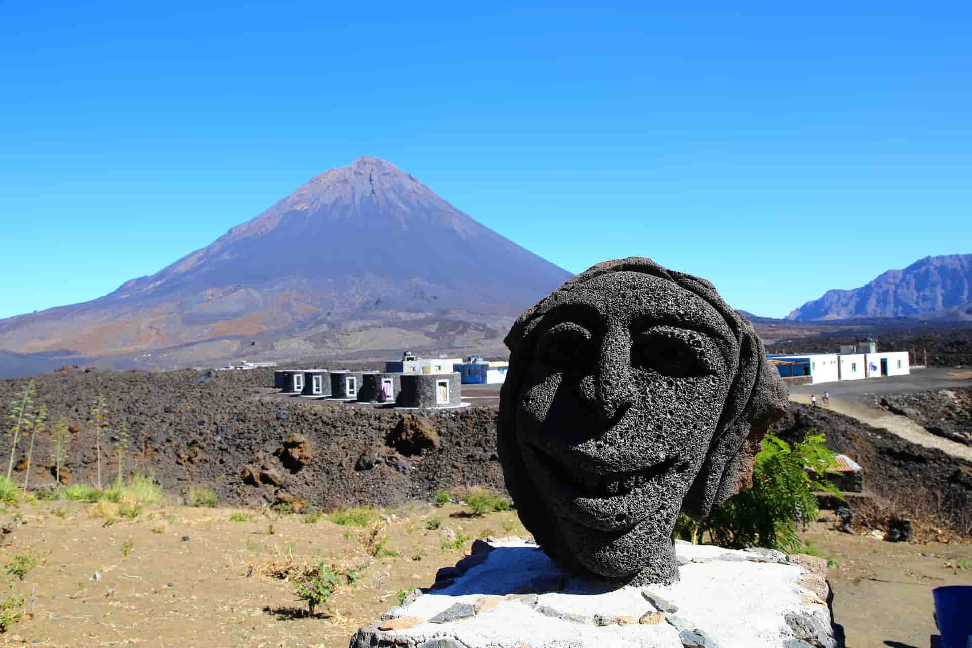 A fascinating close-up view of a smiling face carved from dark volcanic stone at a local settlement on Fogo Island, Cape Verde—a striking volcanic shore excursion destination. The image foregrounds the expressive basalt sculpture, with the sprawling black lava fields of Chã das Caldeiras, modern low-rise stone housing, and the massive, towering cone of the Pico do Fogo volcano rising majestically into a stark blue sky in the background.