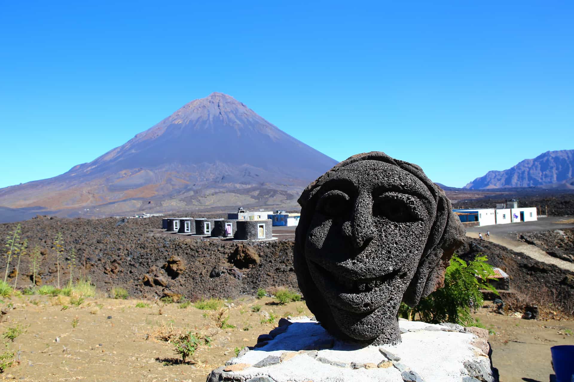 A fascinating close-up view of a smiling face carved from dark volcanic stone at a local settlement on Fogo Island, Cape Verde—a striking volcanic shore excursion destination. The image foregrounds the expressive basalt sculpture, with the sprawling black lava fields of Chã das Caldeiras, modern low-rise stone housing, and the massive, towering cone of the Pico do Fogo volcano rising majestically into a stark blue sky in the background.