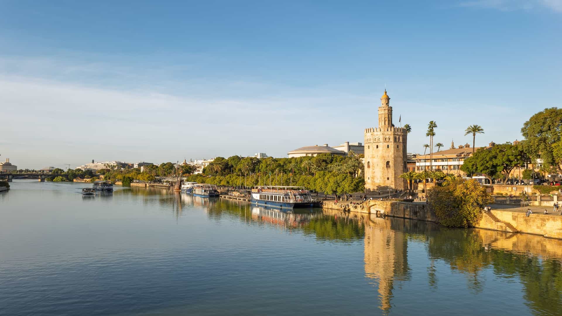 The Torre del Oro overlooking the Guadalquivir River in Seville, with boats moored along the riverside promenade and a clear blue sky above.