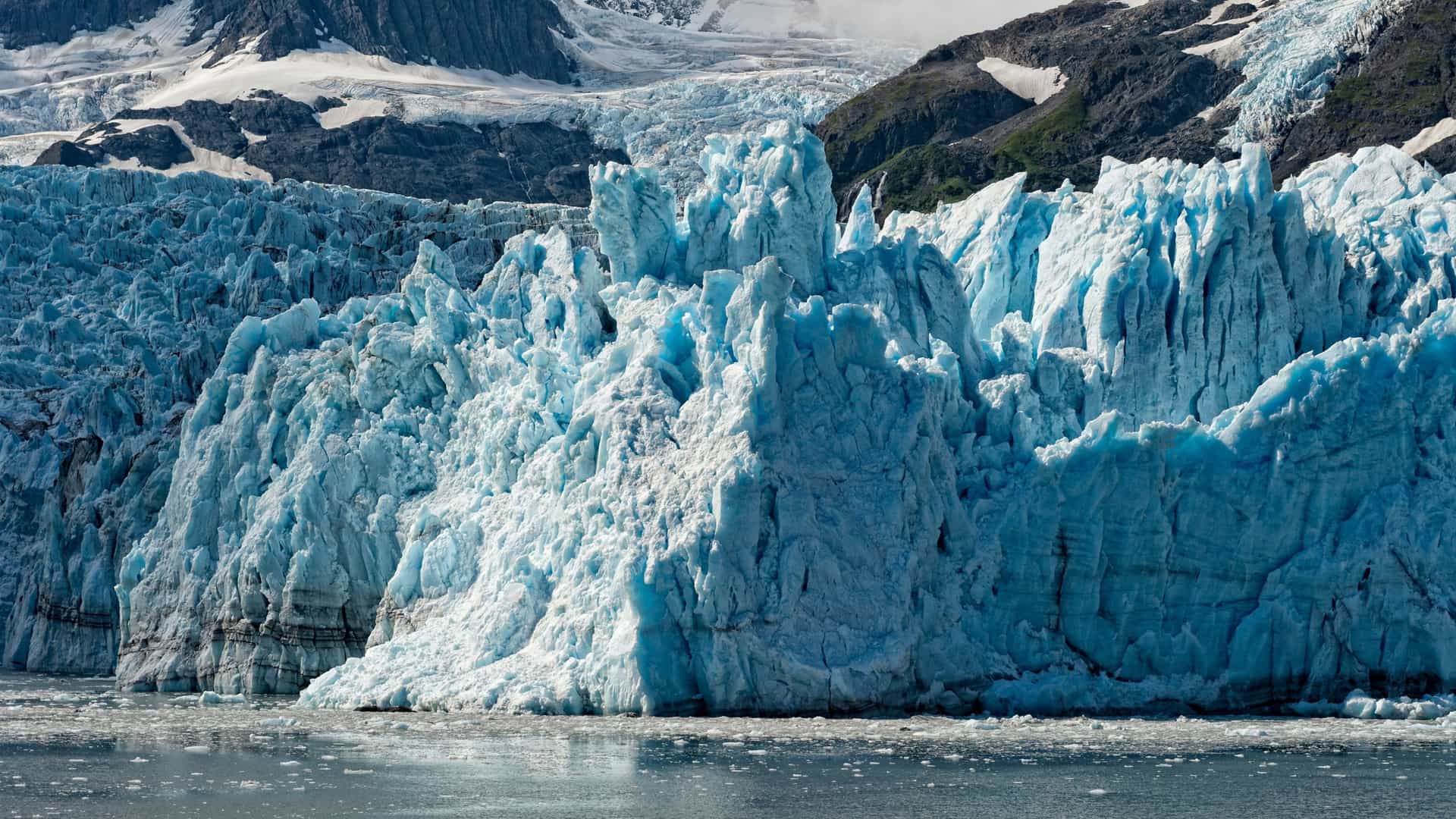A spectacular, close-up landscape view of the jagged, electric-blue ice face of a massive tidewater glacier crumbling into the sea in Harriman Fjord, Alaska, USA—a premier destination for wilderness boat cruises and glacier-viewing excursions. The image highlights the incredibly detailed, towering columns and deep crevasses of the glacial wall meeting dark, ice-filled waters, with steep, snow-streaked mountain ridges rising sharply in the background under a soft light.