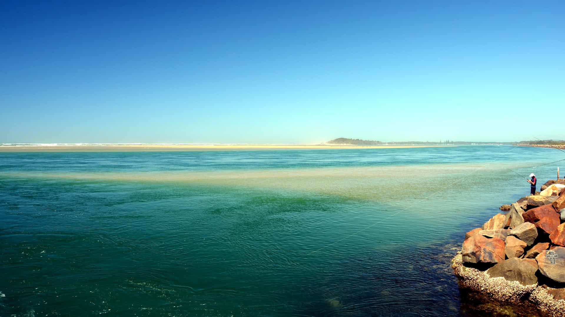 A rocky Harrington Harbour shoreline with deep blue and turquoise water, a lone person fishing on sunlit coastal rocks, and a sandy beach stretching across the horizon under a clear blue sky.