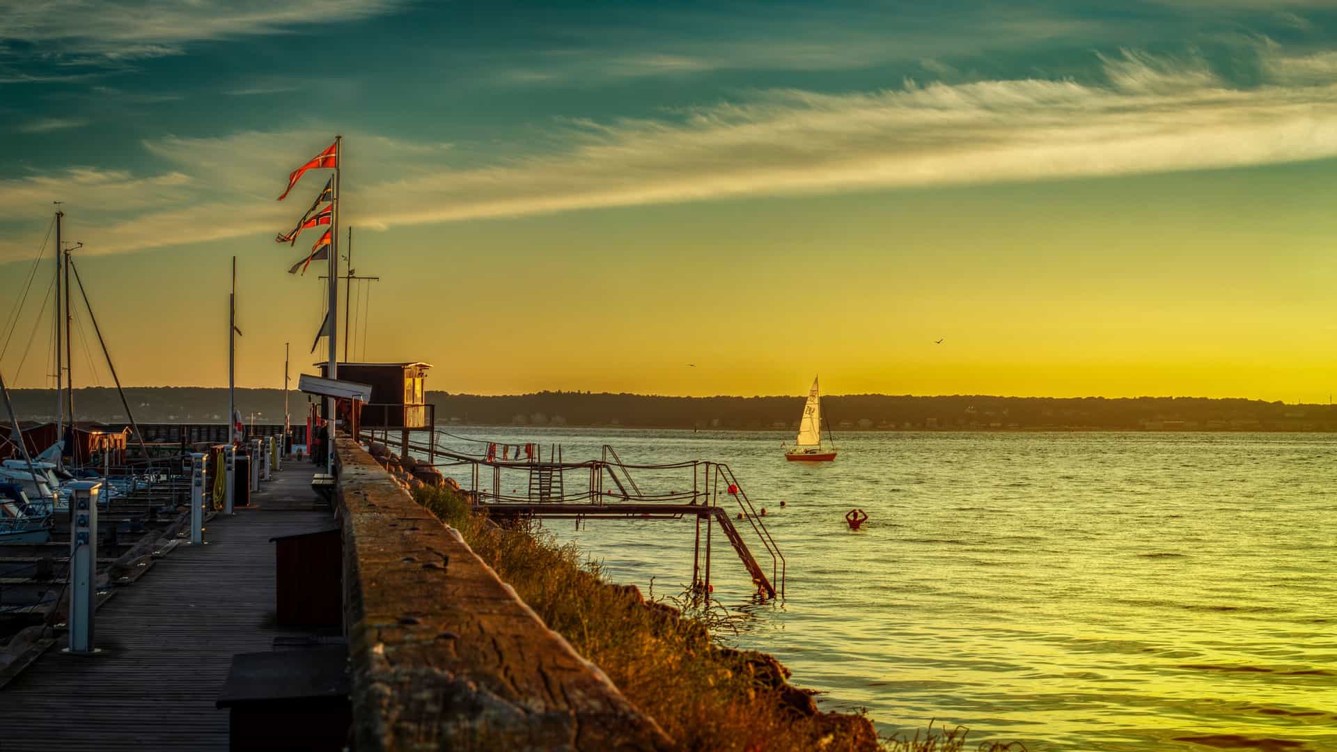 A golden Helsingør sunset over a calm marina, with moored sailboats along a wooden pier, fluttering flags, and a lone sailboat glowing on the Øresund under a blue‑to‑yellow sky.