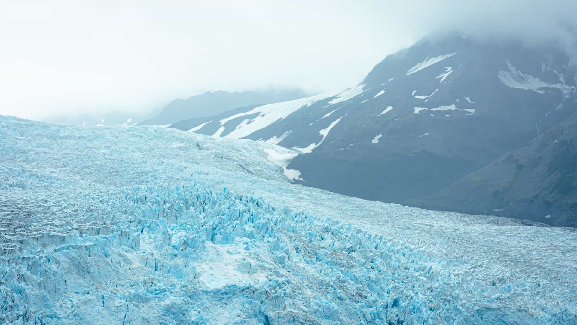 A breathtaking landscape view of a massive, heavily textured tidewater glacier framing a calm, deep-blue bay in Alaska, USA—a premier destination for glacier cruise tours and wilderness excursions (frequently showcased as Holgate Glacier in Kenai Fjords National Park).