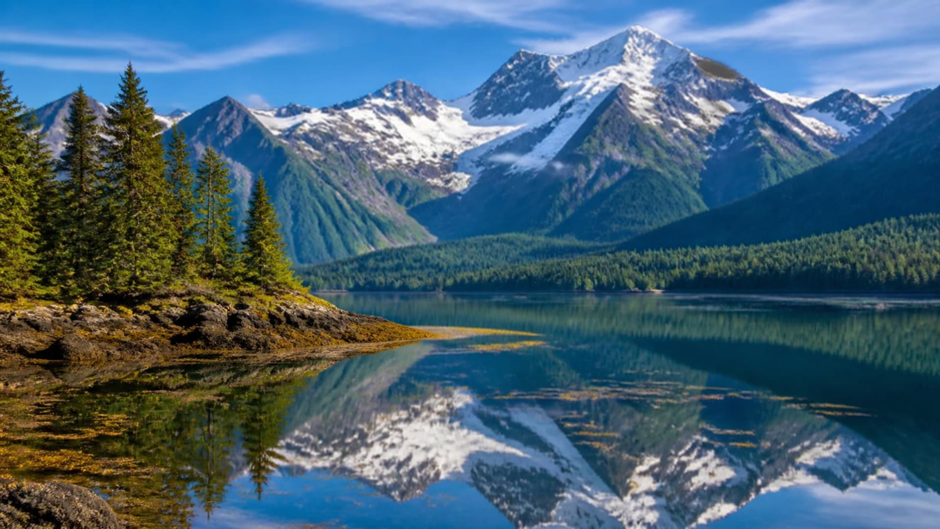 An Alaskan wilderness fjord near Hoonah with snow-capped mountains, dense evergreen forest and calm reflective waters.