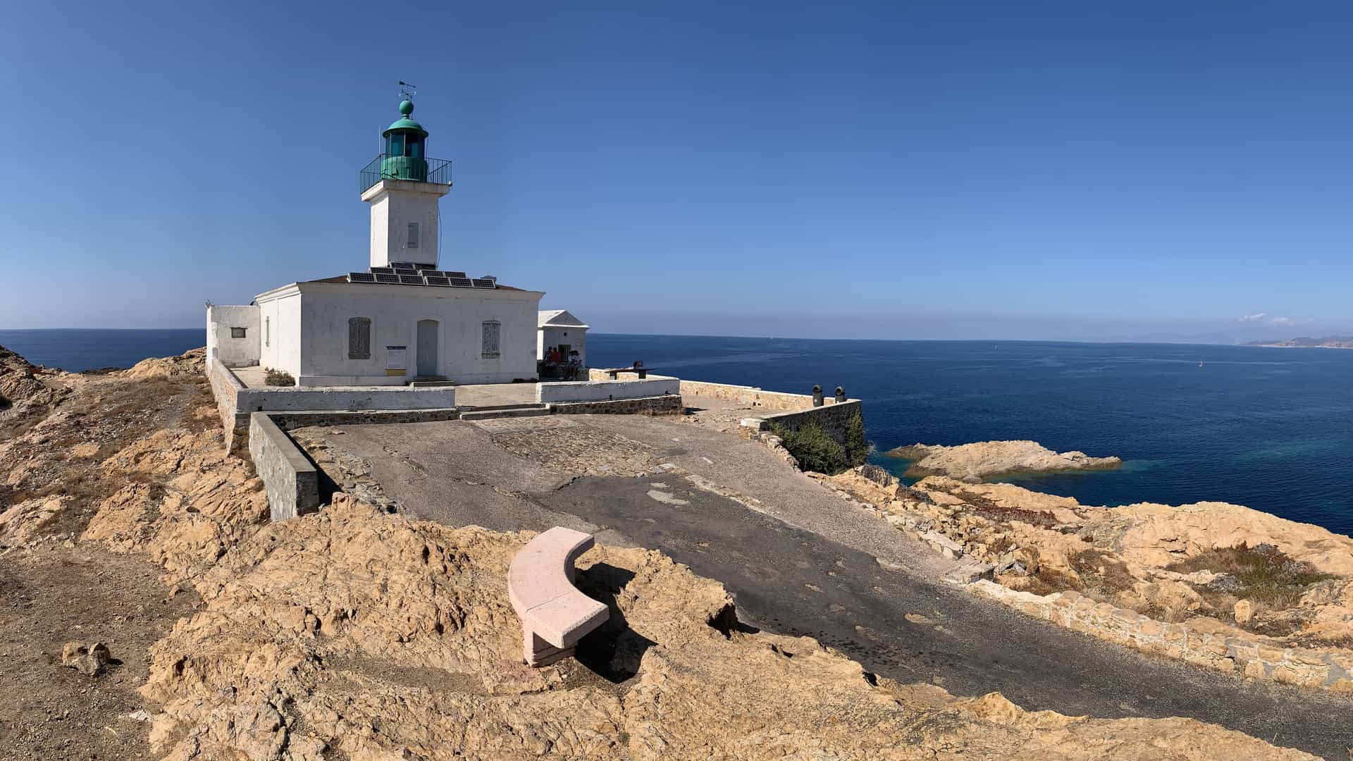White lighthouse and ruined stone tower on red coastal rocks above the harbor of L’Île‑Rousse, Corsica, at sunset