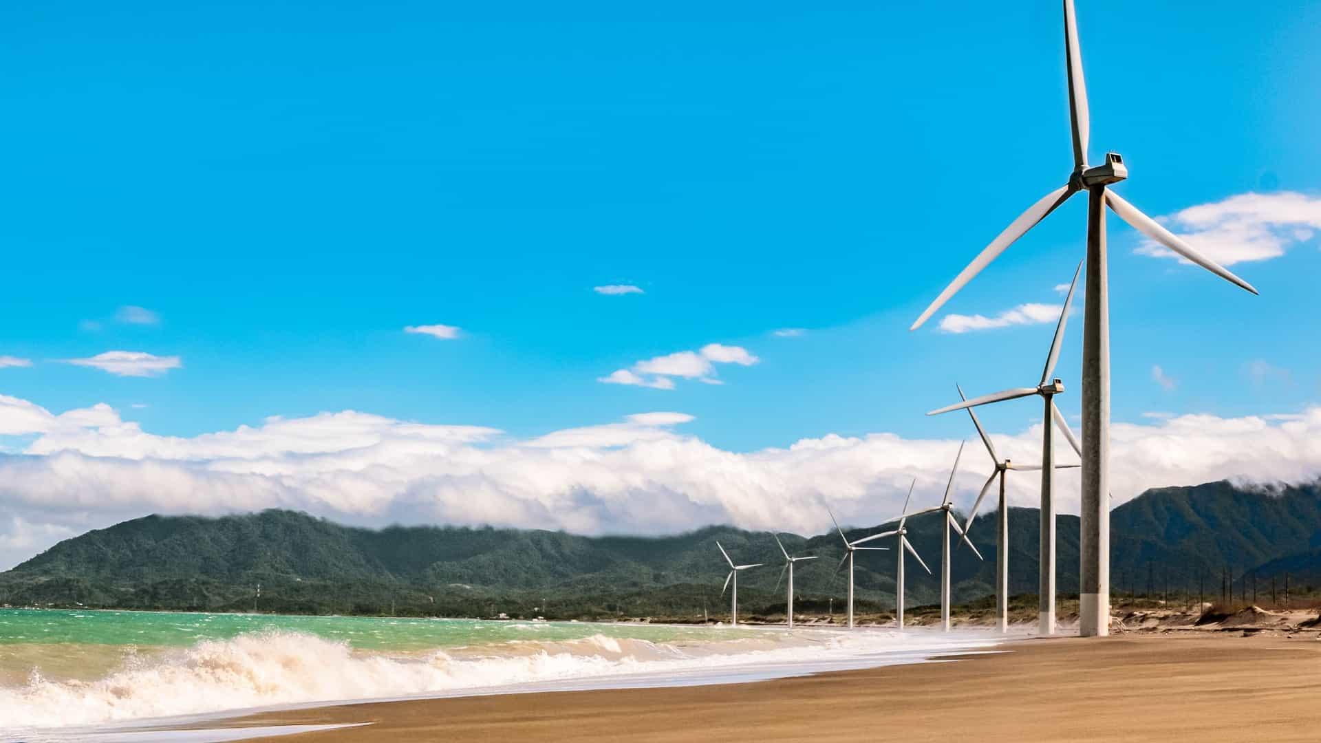 A coastal scene in Ilocos Norte featuring tall wind turbines along a sandy shoreline, waves breaking on the beach, and green mountains rising in the background.