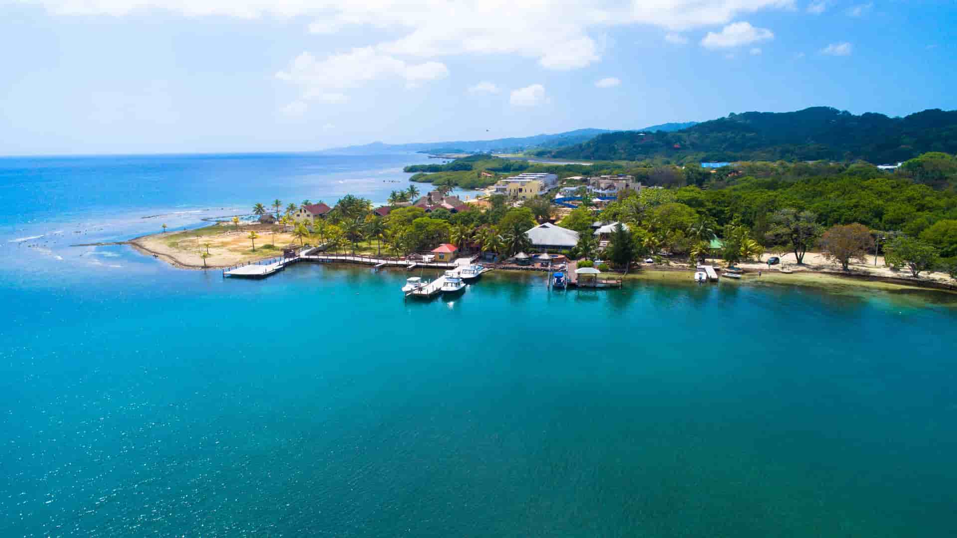 An aerial view of Isla Tropicale showing turquoise water around a small palm‑lined peninsula with docks and boats, sandy beaches, lush green vegetation, and rolling hills stretching along the Honduran coastline under a bright, partly cloudy sky.