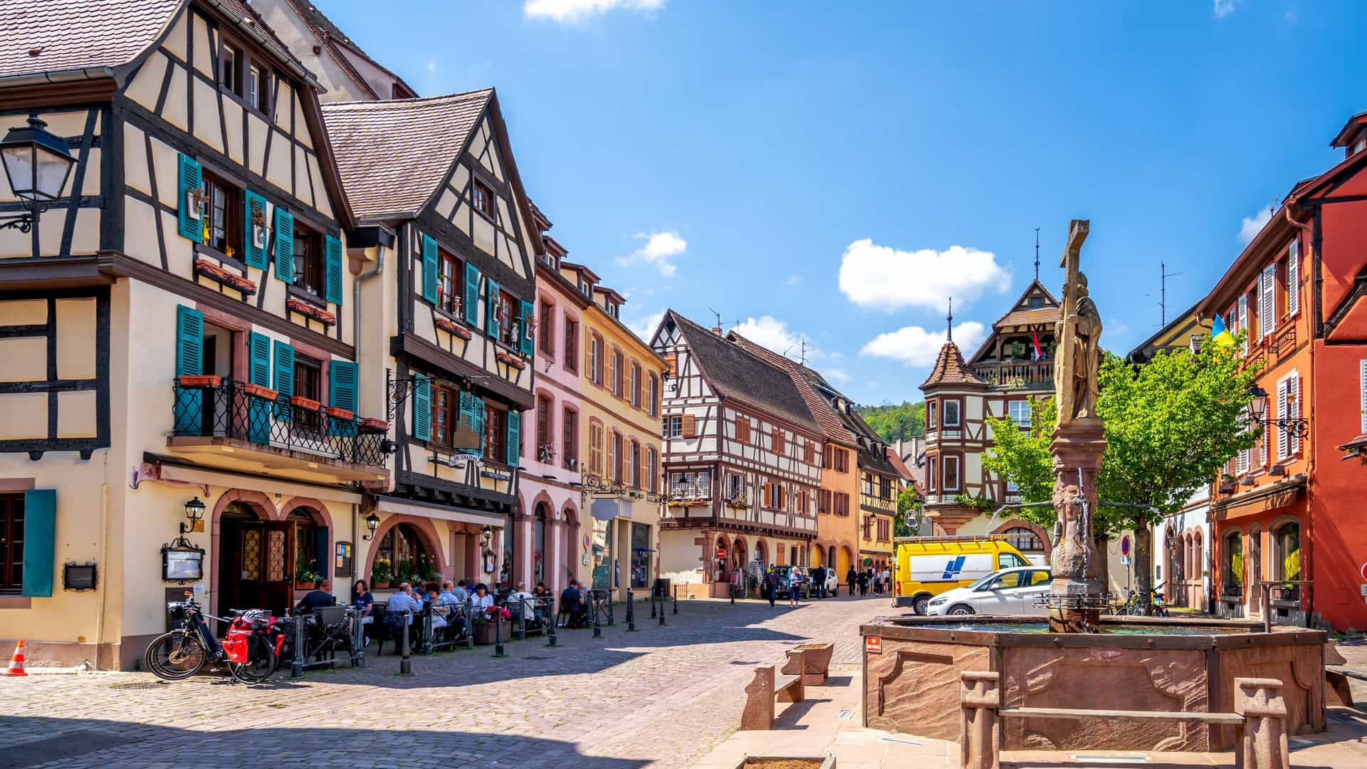 A colorful half‑timbered square in Kaysersberg, France, with pastel façades, a stone fountain, outdoor cafés, and cobblestone streets under a bright sky, showcasing the charm of this Alsace wine‑route excursion stop.