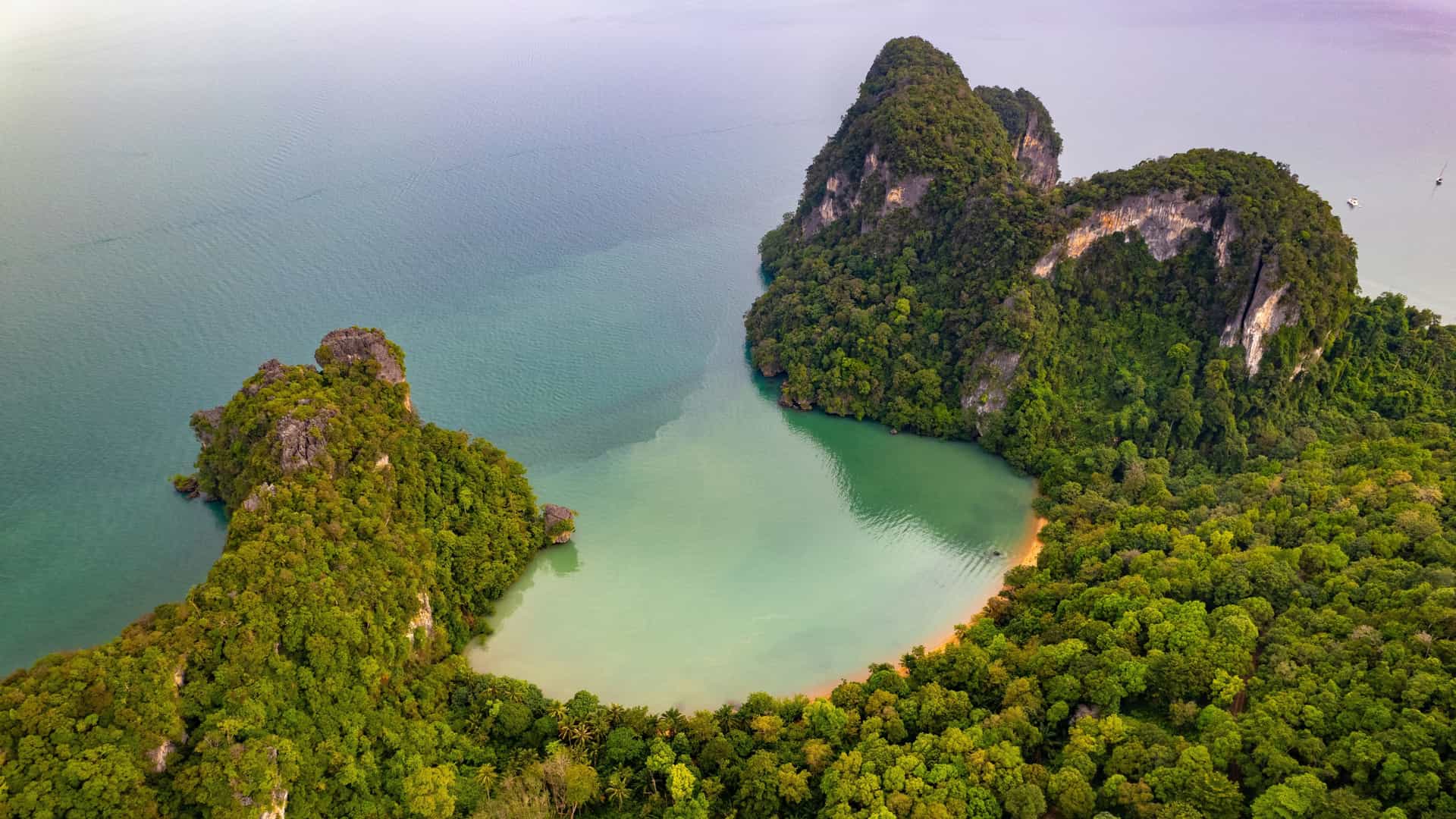 An aerial view of Koh Yao showing steep forested limestone cliffs surrounding a calm turquoise bay, with a narrow sandy beach and lush tropical vegetation along the shoreline under a bright, partly cloudy sky.