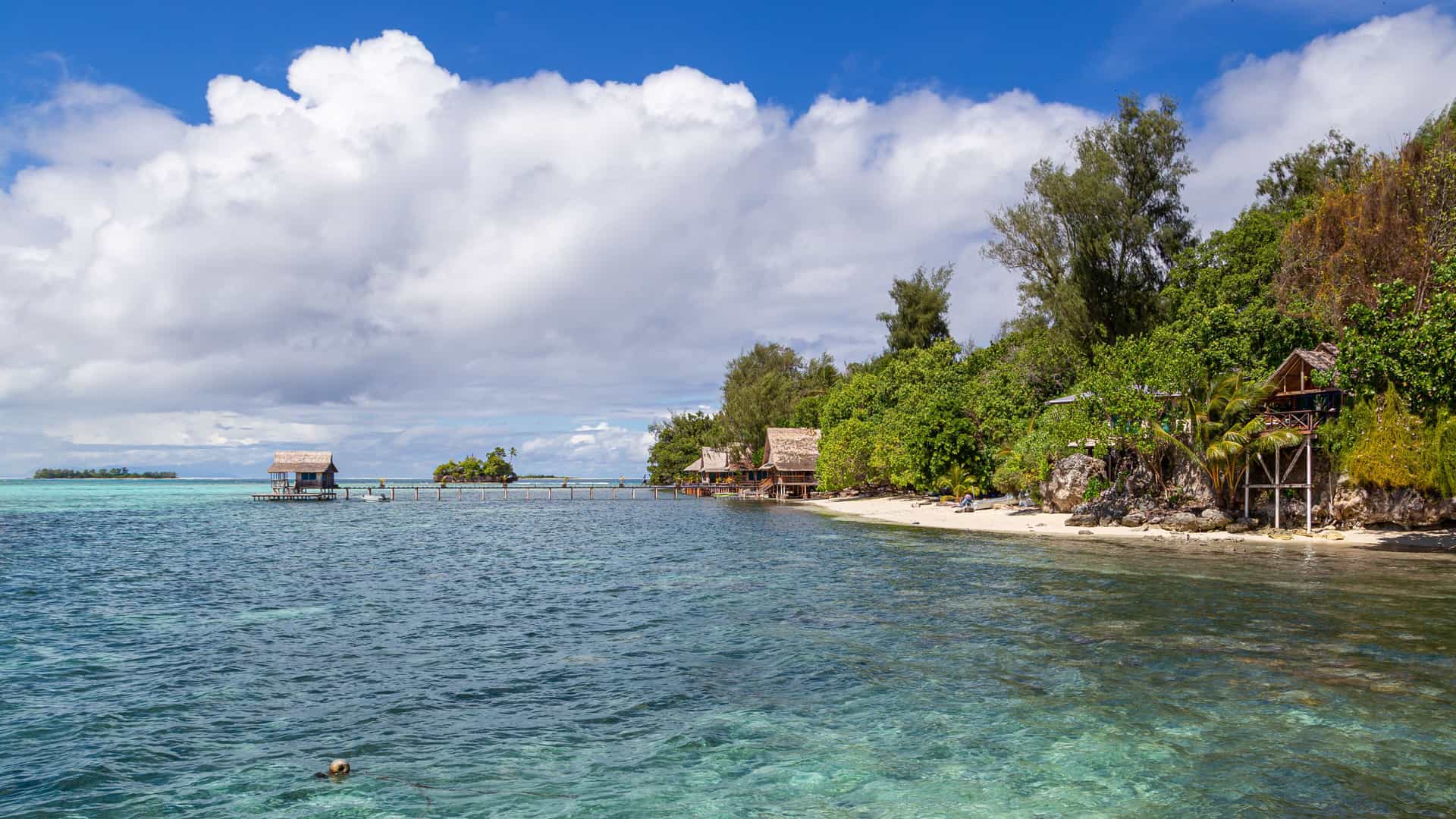 A tropical shoreline on Kolo Hite Island in the Solomon Islands, with turquoise water, sandy beaches, thatched huts on stilts, and lush green vegetation under a bright partly cloudy sky, showcasing the island’s idyllic South Pacific scenery.