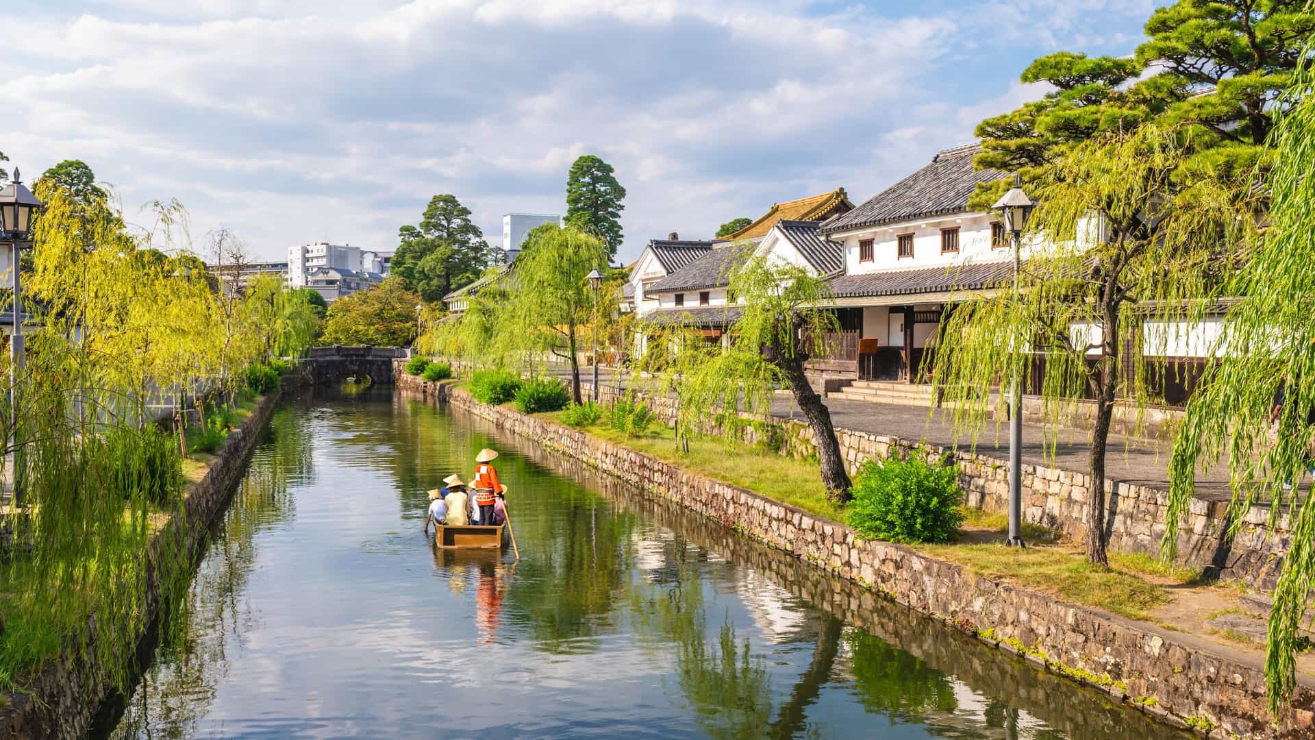 A calm Kurashiki canal lined with willow trees and traditional white‑walled buildings, with a small wooden boat carrying passengers in straw hats across the reflective water.