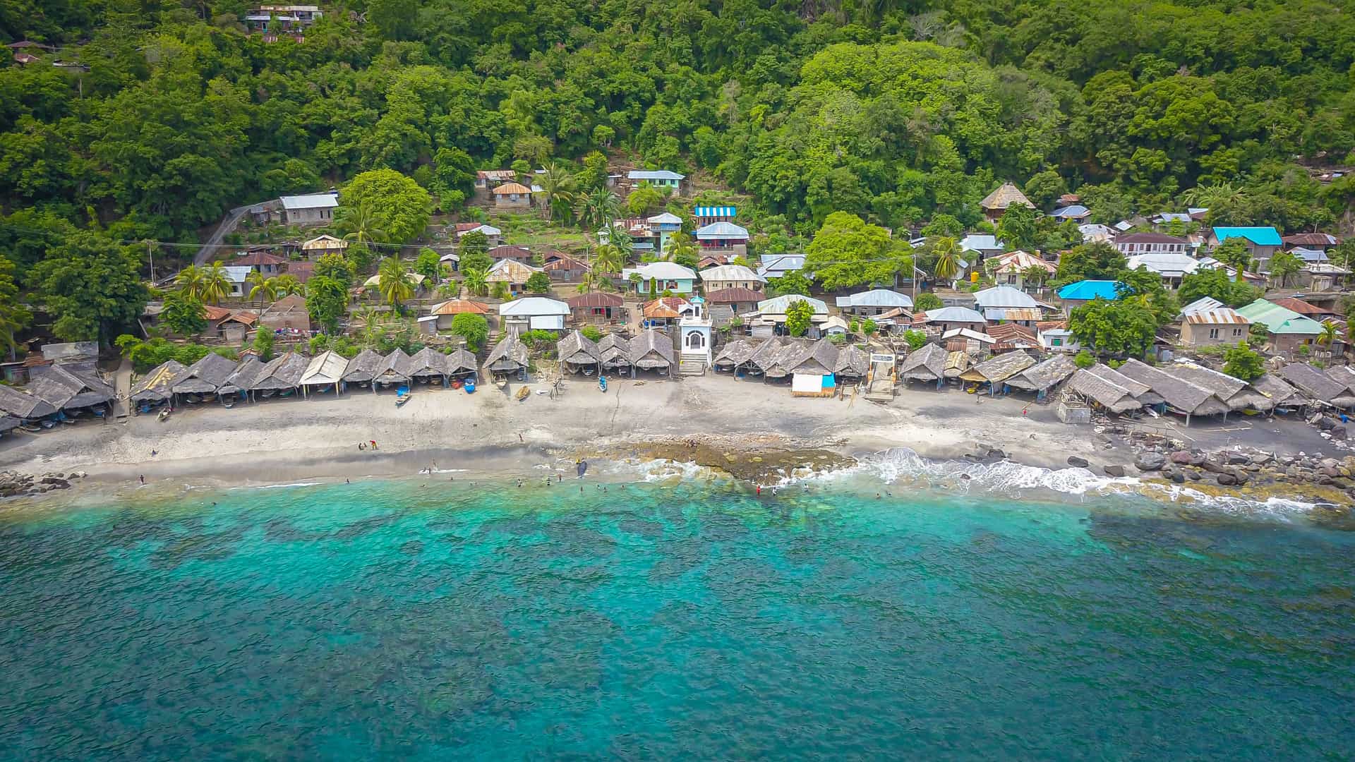 An aerial view of Lamalera village on Lembata Island, showing thatched and metal‑roof homes along a sandy shoreline, rows of coastal huts, turquoise shallows fading into deep blue water, and dense tropical forest surrounding this traditional Indonesian seaside community.