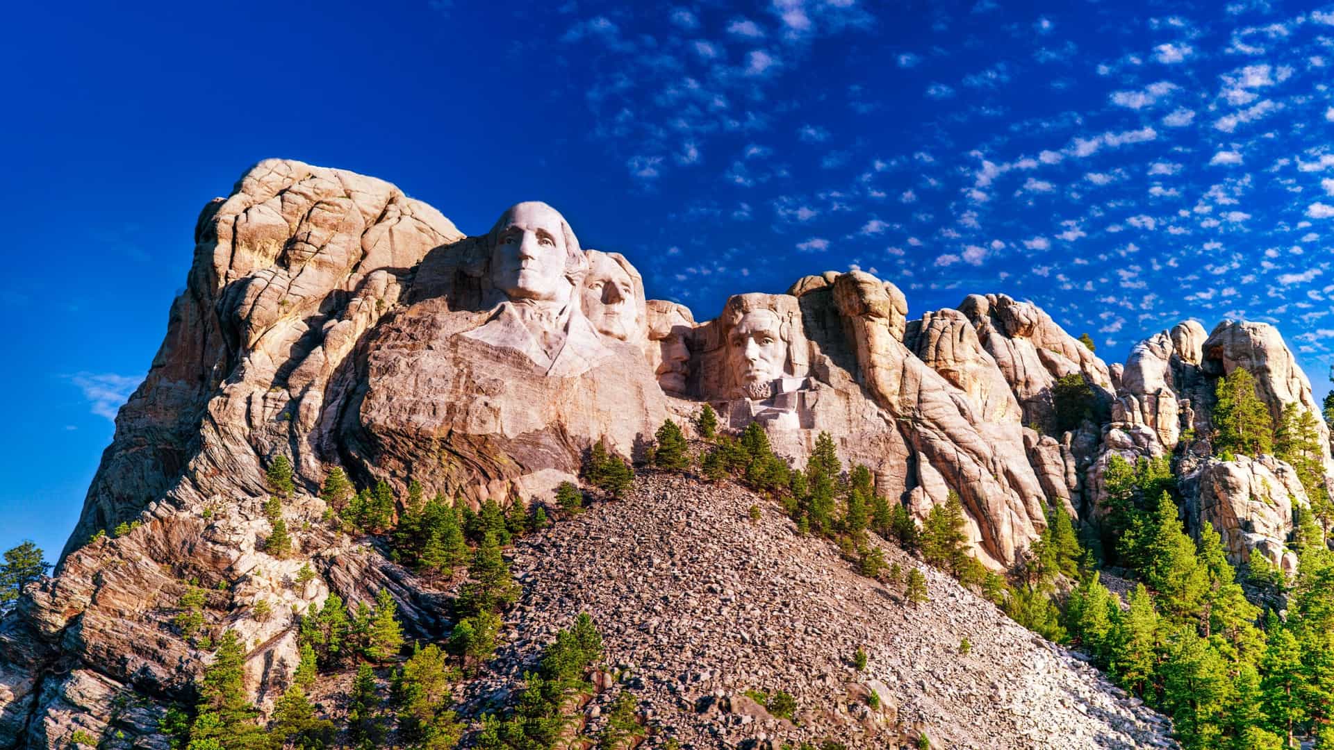 A grand, low-angle landscape view of the Mount Rushmore National Memorial carved into the rugged granite cliffs of the Black Hills in South Dakota, USA—a premier landmark for overland nature tours and cultural excursions. The image showcases the colossal sculpted faces of four American presidents—George Washington, Thomas Jefferson, Theodore Roosevelt, and Abraham Lincoln—glowing under a warm sun, with a steep slope of pine trees and fallen rock debris leading up to the monument against a brilliant blue sky dappled with altocumulus clouds.